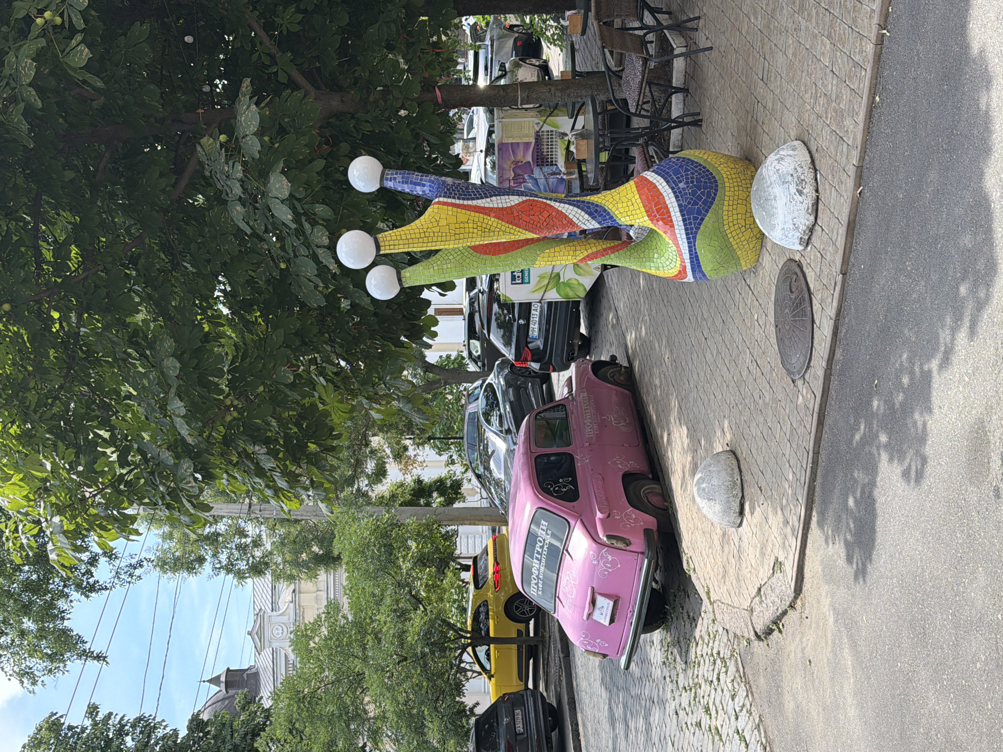 A pink, vintage car is parked on a cobblestone street in Odesa, Ukraine. The car has a license plate and decorative white decals on its sides and rear. Next to the car, a colorful mosaic sculpture stands tall, topped with three white spheres. In the background, other vehicles are parked, and trees line the street. A building with a dome and ornate architectural details is visible in the distance. Outdoor seating with tables and chairs is set up near the sculpture, suggesting a cafe or restaurant. The sky is partly cloudy.