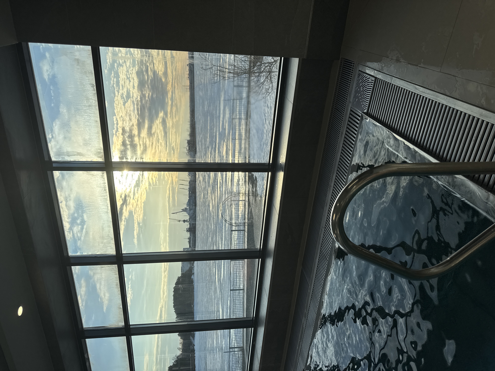 The image depicts a view from inside a building, possibly a spa or swimming pool area. A large window with multiple panes provides a view of a body of water, possibly a river or bay. Across the water, a shoreline is visible, with some industrial structures, including cranes, on the horizon. The sky is cloudy, and the sun is reflecting off the water.
To the right of the window is a section of a pool or tub. Water fills the basin, and a metal grab bar is partially visible in the water. The edge of the pool is lined with a gray grated surface, likely for drainage. The room appears to be tiled with gray tiles.
The location is Chaikovsky, Russia. The body of water is most likely the Votkinsk Reservoir on the Kama River, which is located near Chaikovsky, Russia.