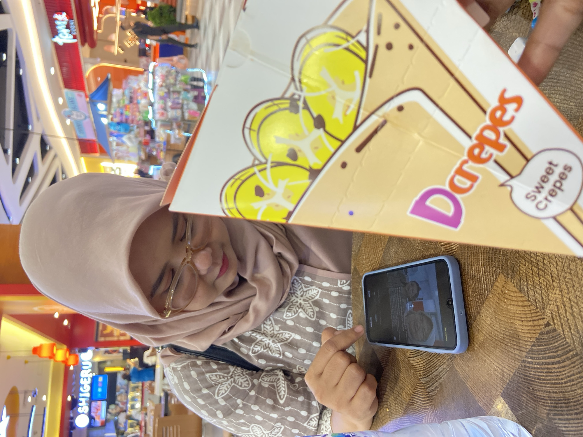 The image shows a woman seated at a table in a brightly lit indoor commercial space, likely a shopping mall or food court in Surabaya, Indonesia. She wears a light brown hijab and eyeglasses, with small, decorative pom-poms attached to the frames. Her attire includes a brown top with a white, intricate floral or star-like pattern.
Her attention is directed downwards towards a smartphone with a light blue case, which rests on the patterned brown table. The phone's screen displays what appears to be a video, possibly a video call, showing two faces. Her right hand, with an extended index finger, is positioned near the phone, suggesting interaction.
Prominently featured in the foreground is a large, triangular cardboard package for "Darepes Sweet Crepes." The packaging is light brown with illustrations of yellow, circular fruit slices, likely bananas, topped with a white substance. The text "Darepes" is visible in orange and purple, along with a speech bubble containing the words "Sweet Crepes." A plastic bottle is partially visible at the bottom of the frame.
The background is blurred, revealing the bustling atmosphere of a mall with various illuminated storefronts. A clear blue sign with white lettering spelling "SHIGERU" is discernible. The woman appears calm and focused on her device. The overall perspective is tilted, giving the scene a dynamic feel.