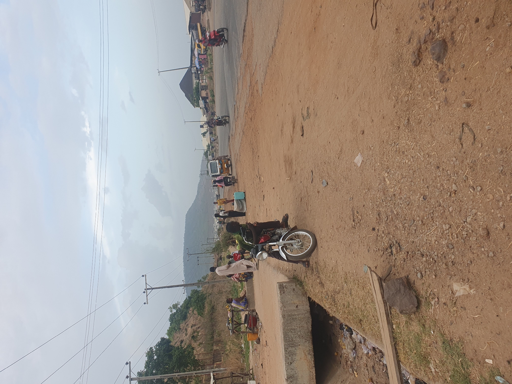 This image depicts a bustling roadside scene, likely in Lokoja, Nigeria, under a hazy sky. The foreground features a dirt path or shoulder alongside a paved road. To the right, a concrete drainage ditch filled with trash and debris runs parallel to the path.
The central subject is a silver and black motorcycle parked on the dirt path. A man wearing dark clothing and a green jacket is seated on the motorcycle, while another man in a light-colored traditional garment stands beside it.
Along the paved road, various activities are taking place. Several individuals are visible, including pedestrians and those gathered near what appear to be market stalls. Some stalls display produce, such as oranges. In the mid-ground, a range of vehicles, including a yellow and white tuk-tuk (keke), other motorcycles, and possibly a car, are present on the road, suggesting ongoing traffic and commercial activity.
Power lines and wooden utility poles crisscross the hazy sky, which has scattered faint clouds. In the distance, a range of hills or mountains forms the backdrop, hinting at the geographical landscape. The overall setting is an everyday street scene characterized by informal commerce, local transportation, and pedestrian movement. There is no discernible text visible in the image. The time of day appears to be daytime, with soft light indicating either morning or late afternoon, or an overcast condition.