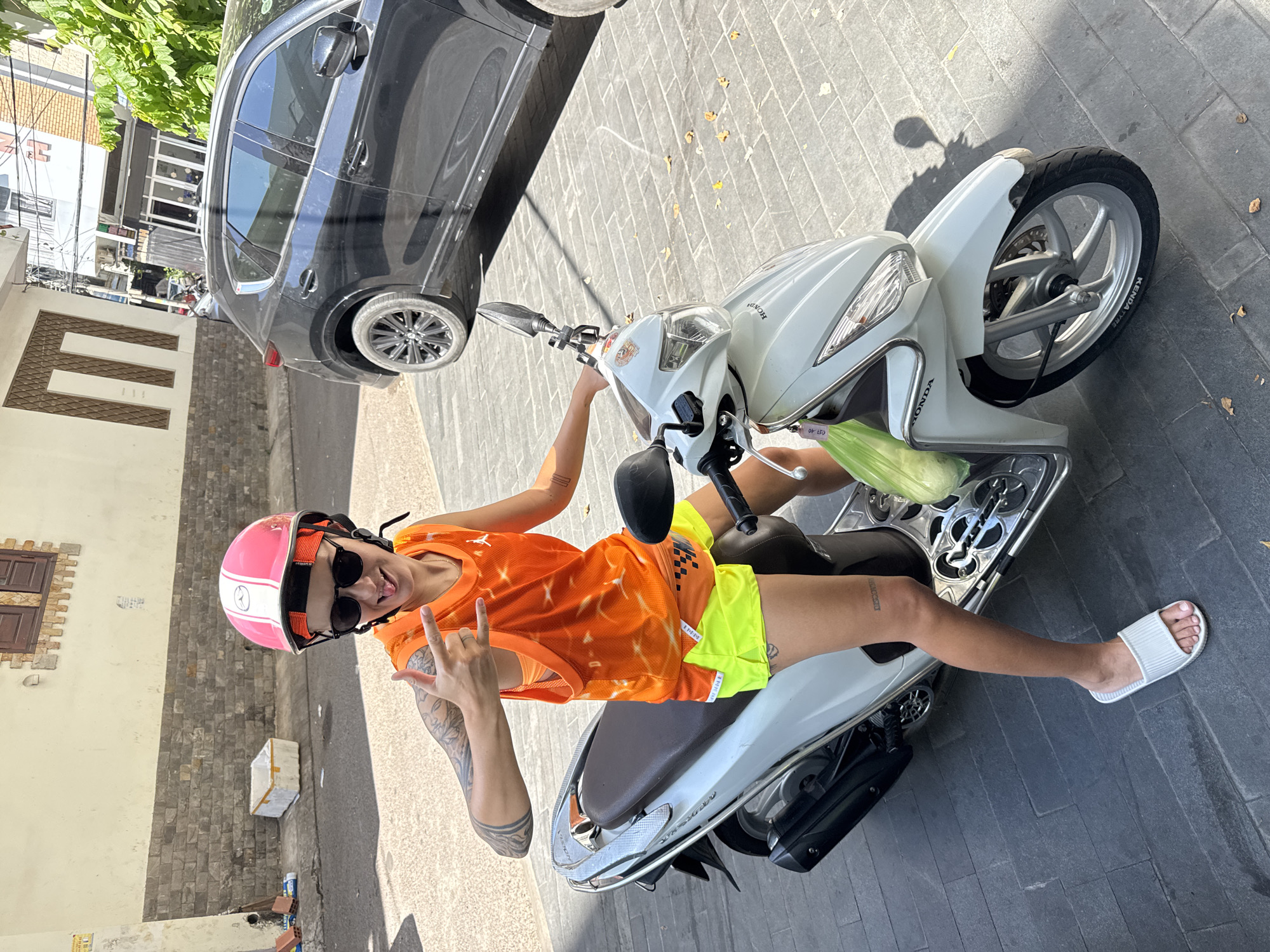 A person is sitting on a white Honda scooter on a paved street in South Quy Nhon Ward, Vietnam. The person is wearing a pink and white helmet, black sunglasses, an orange mesh tank top, and bright yellow shorts. They are sticking out their tongue and making a hand gesture with their right hand. A green plastic bag is hanging from the scooter's handlebars. A black car is parked behind the scooter. The street is paved with dark gray interlocking bricks, and the buildings in the background are made of concrete and brick. Sunlight casts shadows on the street.