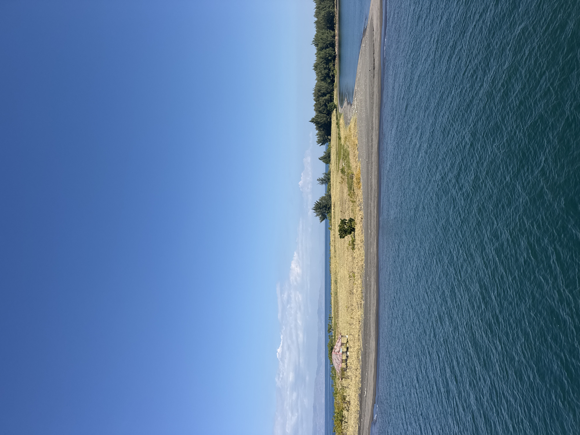 The image shows a tropical landscape with a clear blue sky and calm blue water. In the foreground, the water is a deep blue, with small ripples on the surface. A narrow strip of dark sand separates the water from a grassy, golden-colored field. A small, traditional-style building with a red roof sits in the middle of the field. In the background, a dense line of green trees forms a natural border along the coast. The distant horizon is marked by faint blue mountains. The scene depicts a tranquil and natural environment.