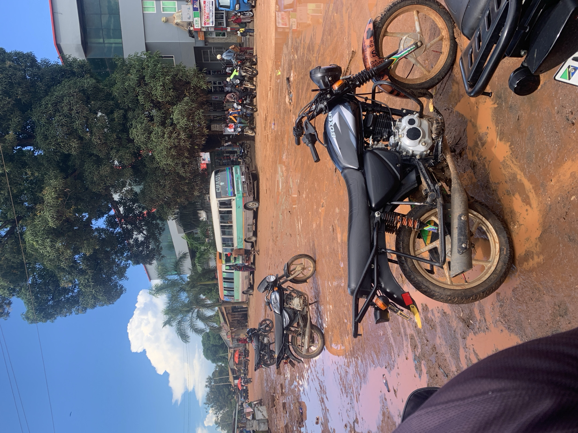 An outdoor scene in Dar es-Salaam, Tanzania, depicts a busy, unpaved urban area saturated with reddish-brown mud and extensive puddles, indicating recent heavy rainfall. The sky is clear blue with prominent white cumulus clouds.
In the immediate foreground, the dark fabric of what appears to be a person's leg or arm is visible in the lower left. Directly in front, the rear section of a black motorcycle is partially obscured. The central focus is a black Bajaj Boxer motorcycle, parked and facing right, displaying custom orange, black, and green accents on its front fender and gold-colored alloy wheels. Its engine casing is silver and the exhaust pipe is mud-splattered. A small green and black graphic, likely part of a Tanzanian flag, is visible near the rear license plate area.
In the midground, another black motorcycle is parked to the left of the central Boxer. Two minibuses are present: one to the left, green and white with "CHAOS" and "KODUVIHOJA VUNGIJA" visible, and another to its right, blue and white with an open passenger door. Numerous individuals, approximately 15-20, are scattered across the scene, walking, standing, and congregating around the vehicles. A cluster of motorcycles and riders is visible to the right of the minibuses. Small stalls with red umbrellas are faintly discernible in the far-left midground.
The background features a multi-story building on the right with a grey facade, green-tinted windows, and red trim at the roofline. Commercial signage, including "WAKALA CUB," is attached. A large tree with dense green foliage partially obscures this building. Palm trees are also visible. To the far left, single-story structures with corrugated iron roofs line the street. Overhead, power lines traverse the scene.