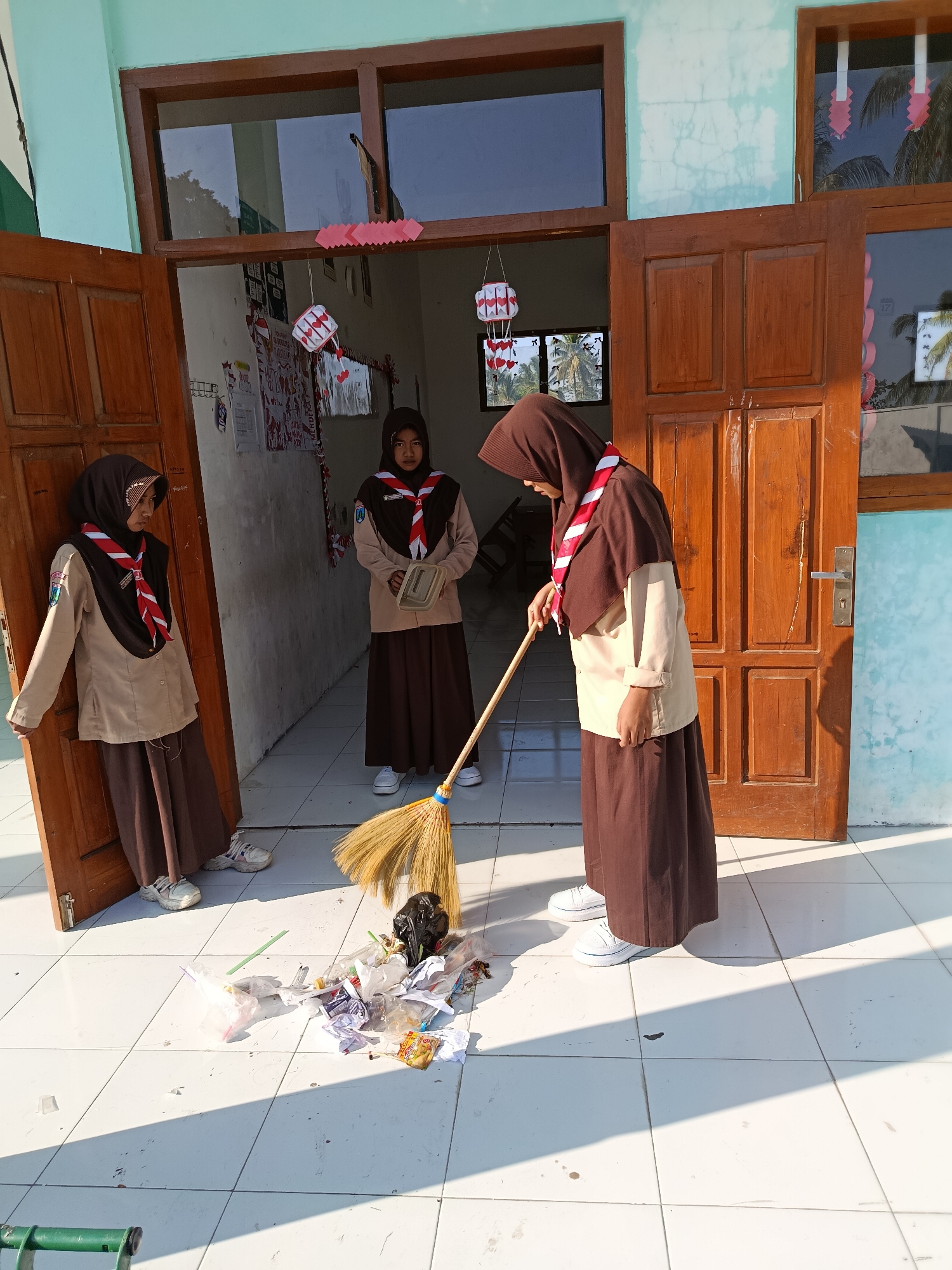 Three young Indonesian girls, dressed in brown and tan scout uniforms with red and white neckerchiefs, are participating in a cleanup activity in front of an open doorway. One girl on the right is actively sweeping a pile of trash on a white tiled floor with a broom. The trash consists of crumpled paper, plastic wrappers, and a black plastic bag. Another girl stands inside the doorway, holding a tan container, and a third girl is leaning against the open door frame on the left. The building appears to be a school, with windows and decorations visible on the walls. The scene takes place outdoors in daylight, with shadows cast on the tiled surface, suggesting a sunny day. The background shows a building facade and glimpses of palm trees through the windows, indicating a tropical or subtropical environment, consistent with the location of Ramban Wetan, Indonesia.