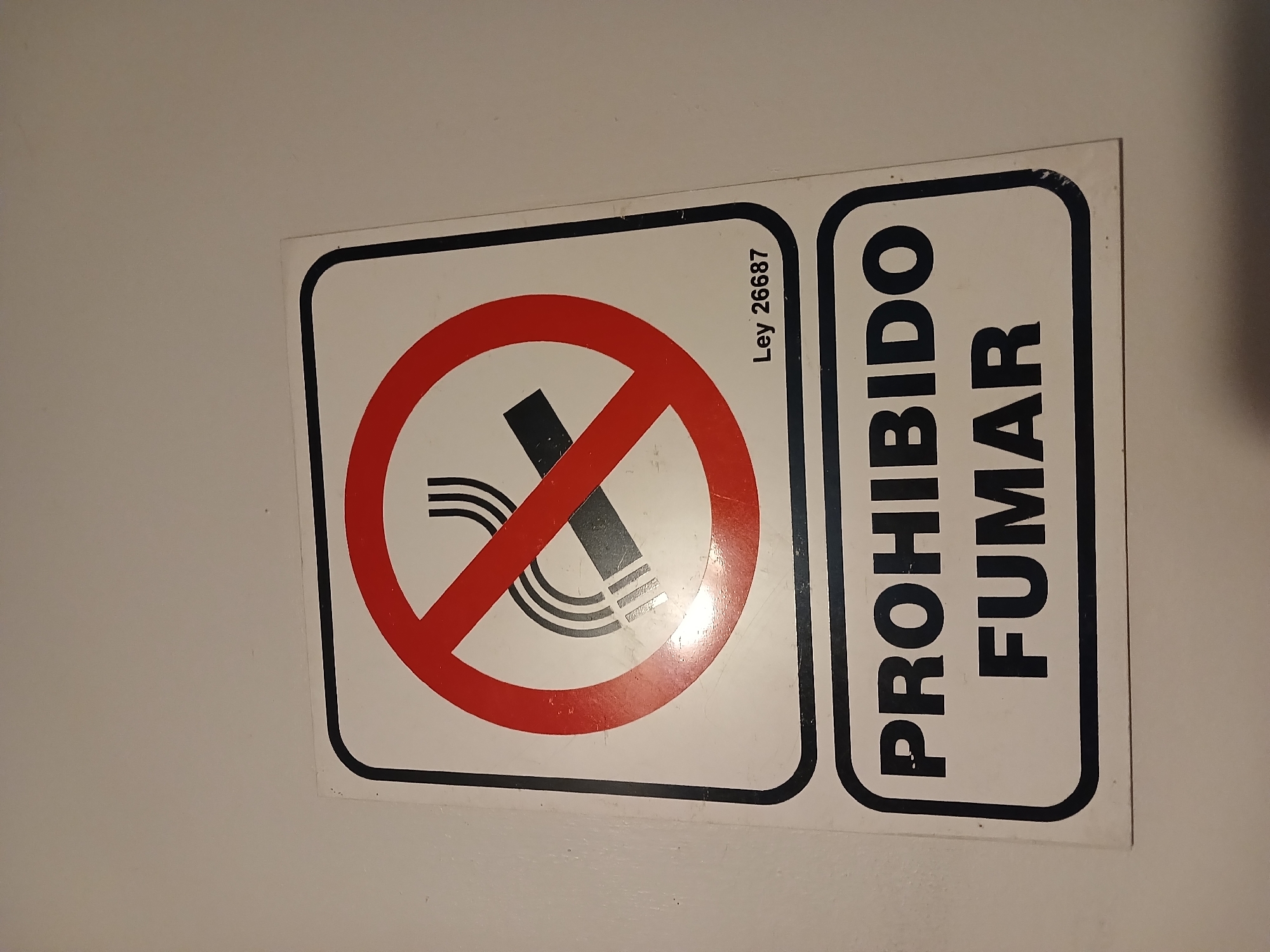 A close-up, slightly angled shot captures a rectangular "No Smoking" sign affixed to a plain, light-colored wall. The sign is divided into two sections. The left section prominently displays the universal "No Smoking" symbol: a black cigarette with three wavy lines indicating smoke, all crossed out by a thick red circle with a diagonal red bar. This symbol is set against a white background, framed by a thin black border.
To the right, the Spanish phrase "PROHIBIDO FUMAR" (NO SMOKING) is printed in large, bold, black capital letters, oriented vertically on a white background, also with a black border. A smaller, vertically aligned text, "Ley 26687," is visible within the white area of the symbol section, referring to an Argentine law related to tobacco product regulation. The sign appears to be a decal or sticker, showing minor surface wear and reflections from artificial lighting. This sign serves as a regulatory directive in an indoor setting within Buenos Aires, Argentina, indicating a smoke-free environment in accordance with Law 26687.