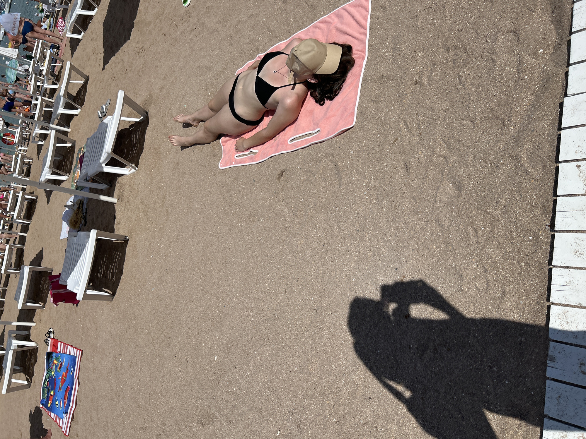 A woman is sunbathing on a pink towel on a sandy beach. She is wearing a black bikini and a beige baseball cap. There are several white lounge chairs and beach towels visible in the background. The scene is a beach in Gelendzhik, Russia, on a sunny day.
