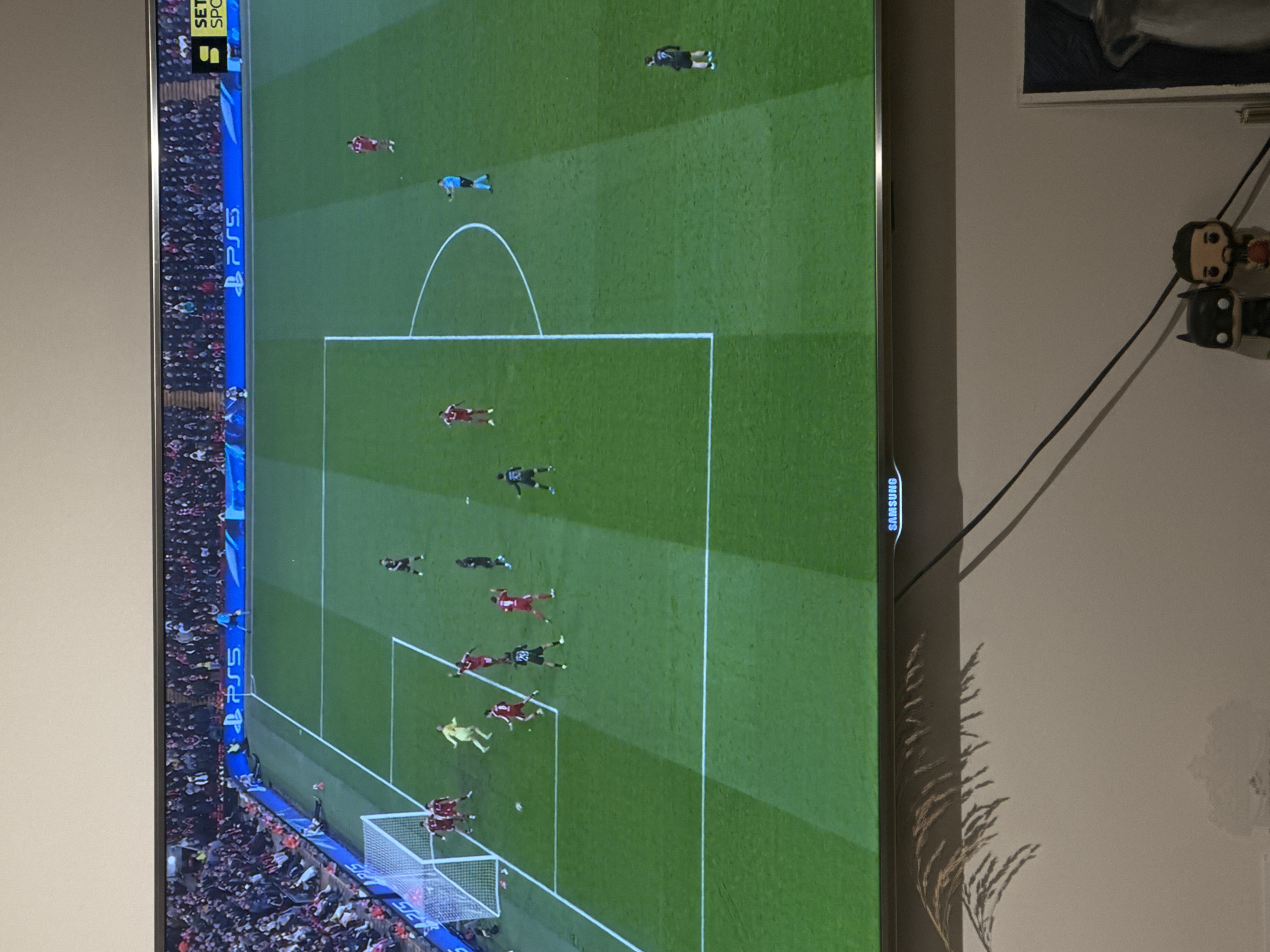 The image shows a television screen displaying a soccer game. The game is being played on a green field marked with white lines. The field is populated with players wearing different colored uniforms, including red and black. Spectators fill the stands along the sidelines. A 'PS5' logo is visible along the edge of the field. The television itself is a Samsung model, positioned against a white wall. Cables are connected to the television. Two figurines are mounted on the wall next to the television.
