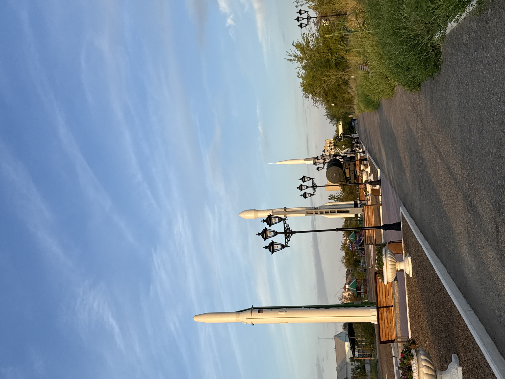 A paved walkway lined with streetlights, planters, and park benches extends into the distance. On the left, three rockets stand vertically. The largest rocket, painted white, has a green stripe running down its side. To its right, two smaller rockets are also visible. In the background, there's a playground with colorful equipment and some buildings. A train car is also parked near the rockets. The sky is a clear blue with wispy clouds. The scene appears to be a park or memorial area in Baikonur, Kazakhstan.