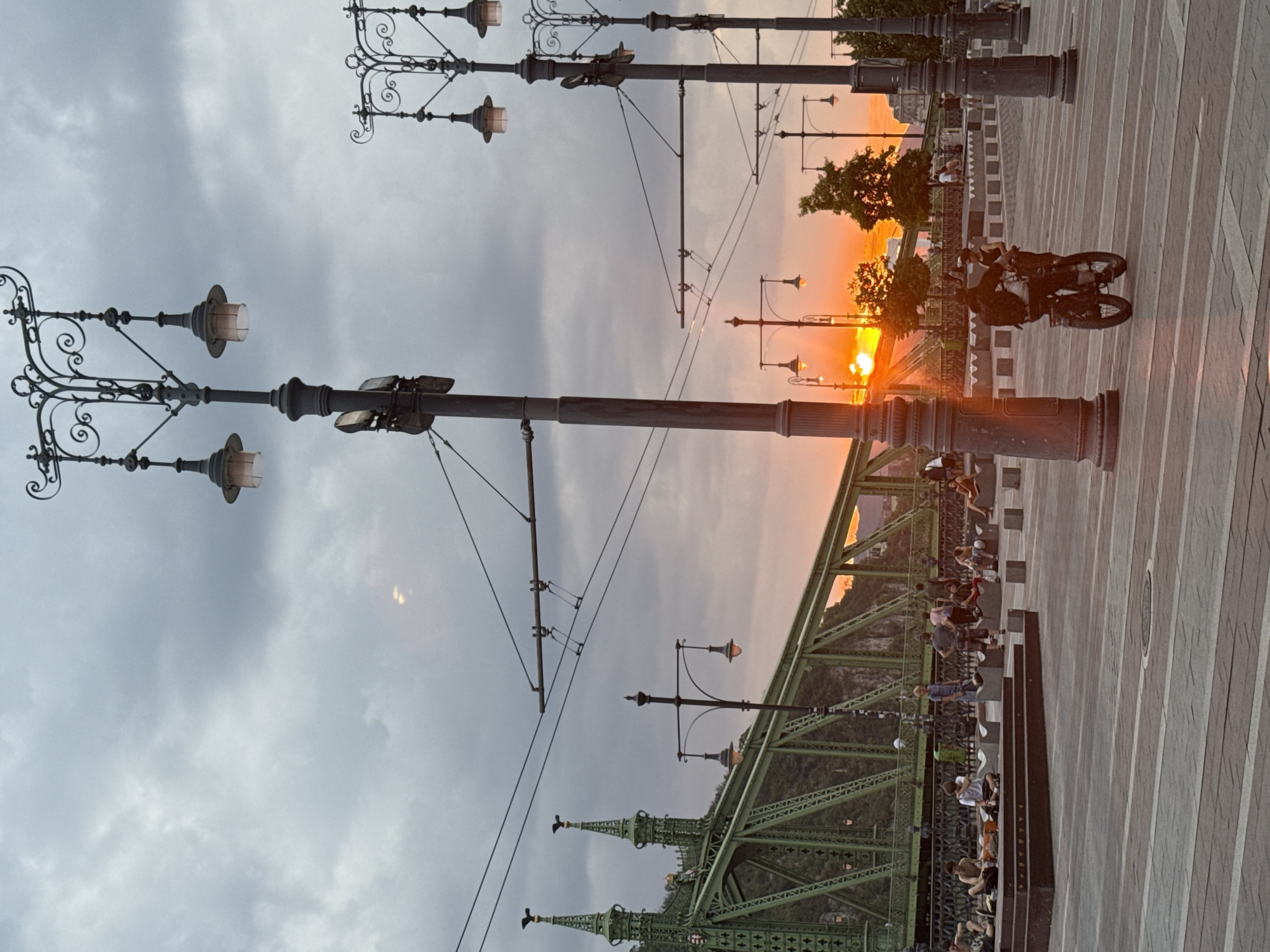 In Budapest, Hungary, at dusk, Liberty Bridge, a green truss bridge, spans the Danube River under a cloudy sky with the setting sun visible. Several streetlights with ornate fixtures line a paved pedestrian area in the foreground. People are gathered on benches and steps near the bridge, while two individuals ride a bicycle on the plaza. The scene captures a moment of leisure and urban life against the backdrop of a dramatic sunset.