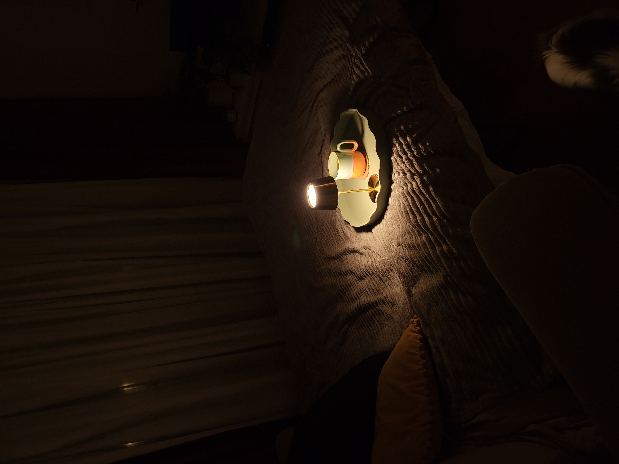 A dimly lit interior features a wall-mounted lamp with a cylindrical light projecting light. The lamp has a cup-like design mounted on a green, asymmetrical base. The wall is covered in textured material. Below the lamp, an orange pillow leans against a larger, possibly sofa, form. The image shows a room, perhaps a bedroom, that is darkened. No location is specified in the image. No people are visible.