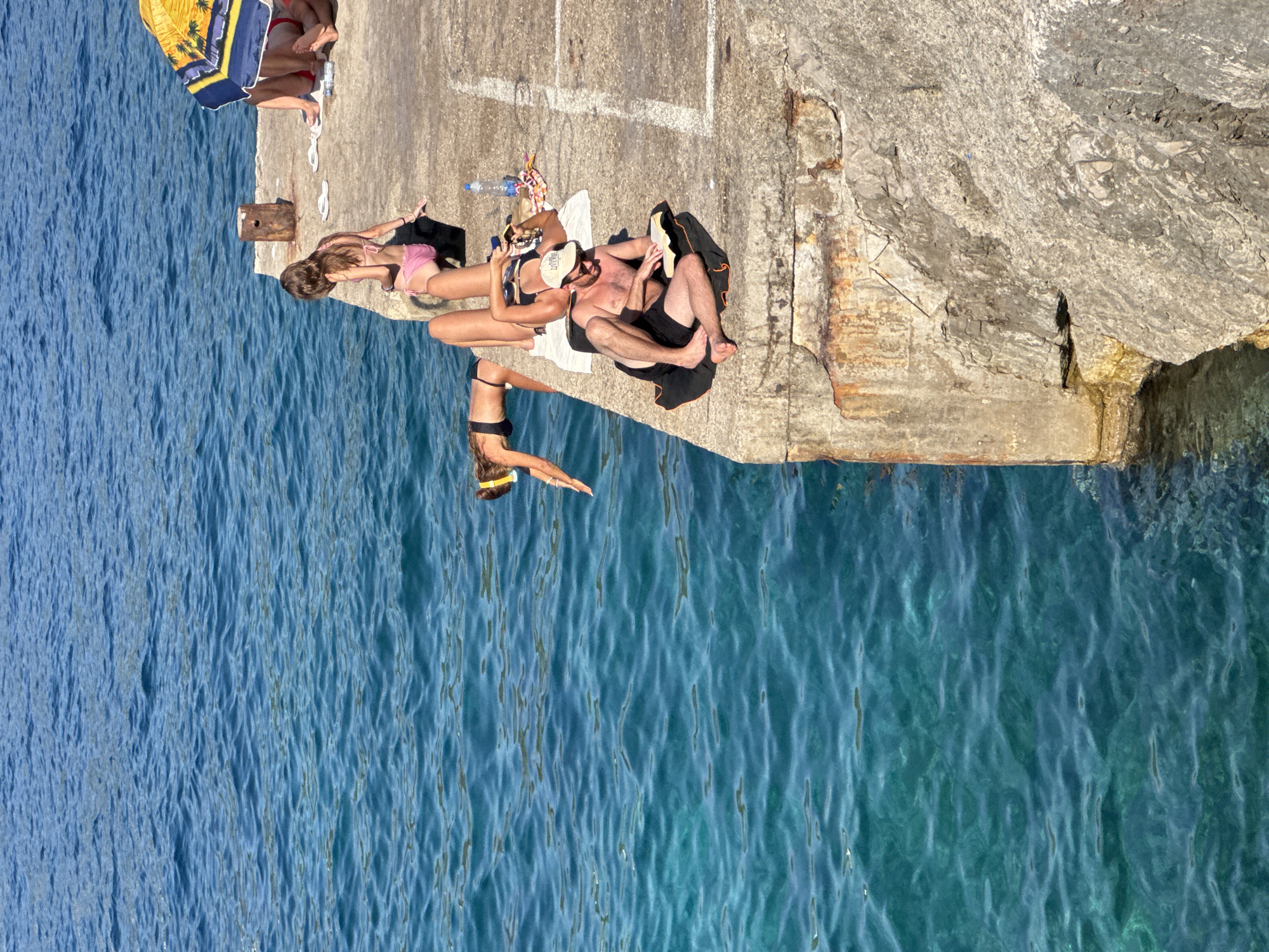 The image depicts a sun-drenched day at a seaside location, likely in Dubraljevina, Montenegro, with people relaxing and preparing to swim. The scene is set on a concrete pier adjacent to clear blue-green water.
In the foreground, a man wearing a light-colored baseball cap and dark shorts is seated on a dark towel, engrossed in reading a book. He is positioned on the edge of the pier, with his legs extended. Next to him, a woman in a black bikini is lying on her back on a white towel, holding a smartphone. A clear plastic water bottle and a colorful beach towel are also visible nearby.
Further along the pier, a young woman in a pink bikini is seated, facing the water. To her left, another woman in a black bikini and a yellow snorkel mask is leaning forward, appearing to be preparing to enter the water. Her body is angled towards the sea, with her hands extended. In the background, partially obscured by a yellow and blue umbrella, are the legs of another person, suggesting more individuals are present.
The environment is characterized by the vibrant blue of the sea, the rough texture of the concrete pier, and the bright sunlight casting shadows. A weathered wooden post stands near the center of the pier. The overall atmosphere suggests a leisurely summer day by the coast.