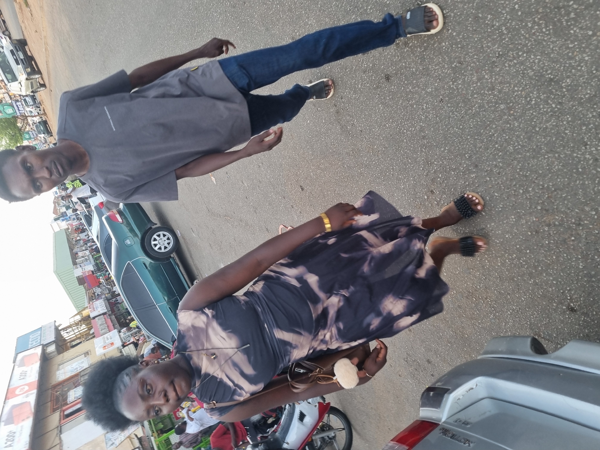 The image shows two individuals, a man and a woman, on a street in Jos, Nigeria. The man is wearing a grey t-shirt and blue jeans, and sandals. The woman is wearing a tie-dye dress and sandals. There are cars, motorcycles, and buildings in the background. The street appears to be paved. The scene is in daylight.