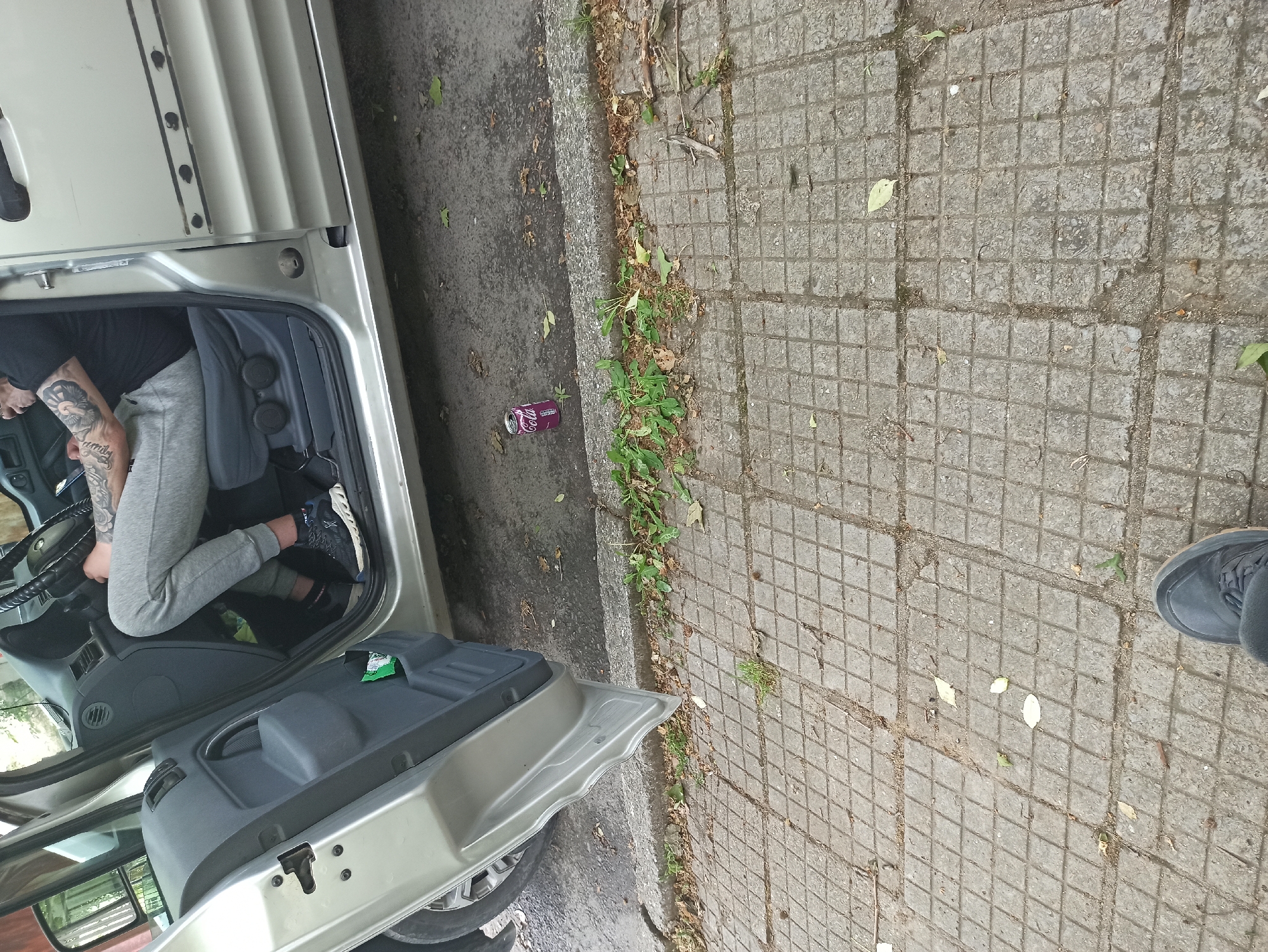 A person is inside a vehicle, with their upper body in the driver's seat, legs extending towards the passenger side, wearing black shirts, gray pants, and black and gray sneakers. The vehicle door is open. Next to the vehicle, on a paved surface, there is a discarded purple soda can. A portion of a shoe is visible on the right side of the image. The background consists of concrete slabs and roadside vegetation.