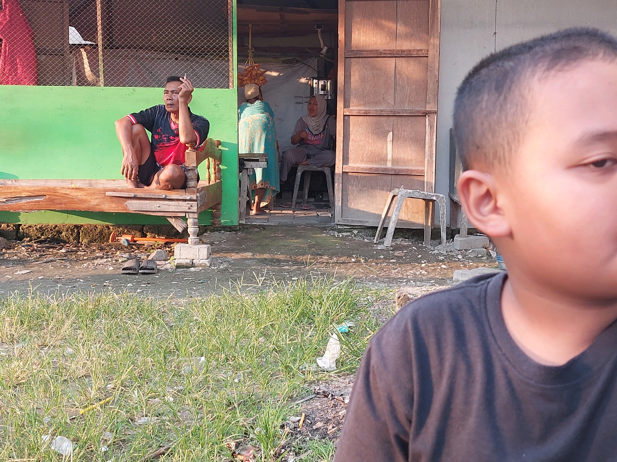 In Canga'an, Indonesia, a man sits on a wooden bench outside a building. He is smoking a cigarette, holding it between his fingers and bringing it to his mouth. He is wearing a dark t-shirt with red accents and dark shorts. To the left of the bench, a green wall is visible, and behind it, a wire fence. Inside the building, two women are present. One woman, wearing a light blue patterned fabric and a headscarf, stands facing away from the camera. The second woman sits on a stool in front of a doorway, wearing a grey t-shirt and a light-colored headscarf, and appears to be speaking or gesturing. The foreground shows a patch of dry grass with some scattered debris. In the immediate foreground, the shoulder and side of a young boy's head are visible, suggesting he is positioned very close to the camera, looking towards the scene. The environment appears to be an outdoor area with a rustic building and a mix of grassy and dirt ground.