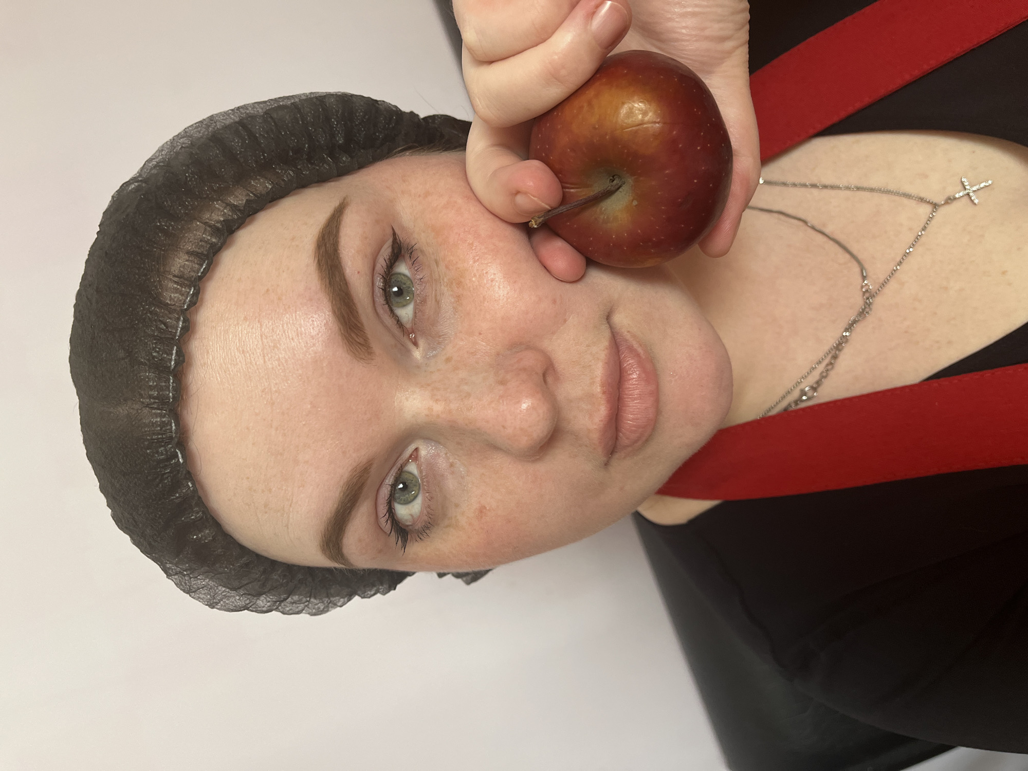 A young woman wearing a black hairnet is holding a red apple up to her face. She is wearing a silver chain necklace with a cross pendant. A red strap can be seen across her shoulder. The background appears to be a plain white surface. The location of the image is not possible to determine.