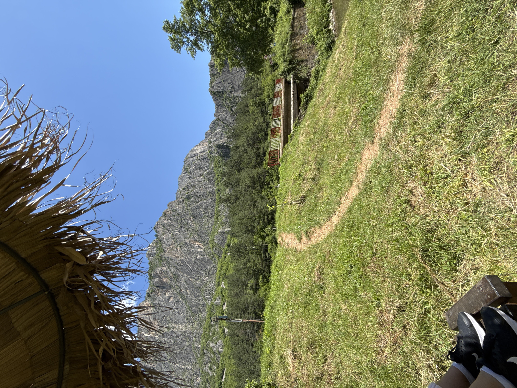 The image depicts a scenic outdoor environment with a prominent rocky mountain in the background. In the foreground, there is a patch of green grass with a dirt path winding through it. A portion of a thatched umbrella is visible in the upper left corner. Further in the background, a forest of green trees lines the base of the mountain. A bridge with red and white railings spans a body of water, and to the right of the bridge, there is a large green tree. A small part of a person's legs wearing black athletic shoes and white socks is visible in the lower left corner, seated on what appears to be a wooden bench. The sky is clear and blue. The location is identified as Zgorigrad, Bulgaria.