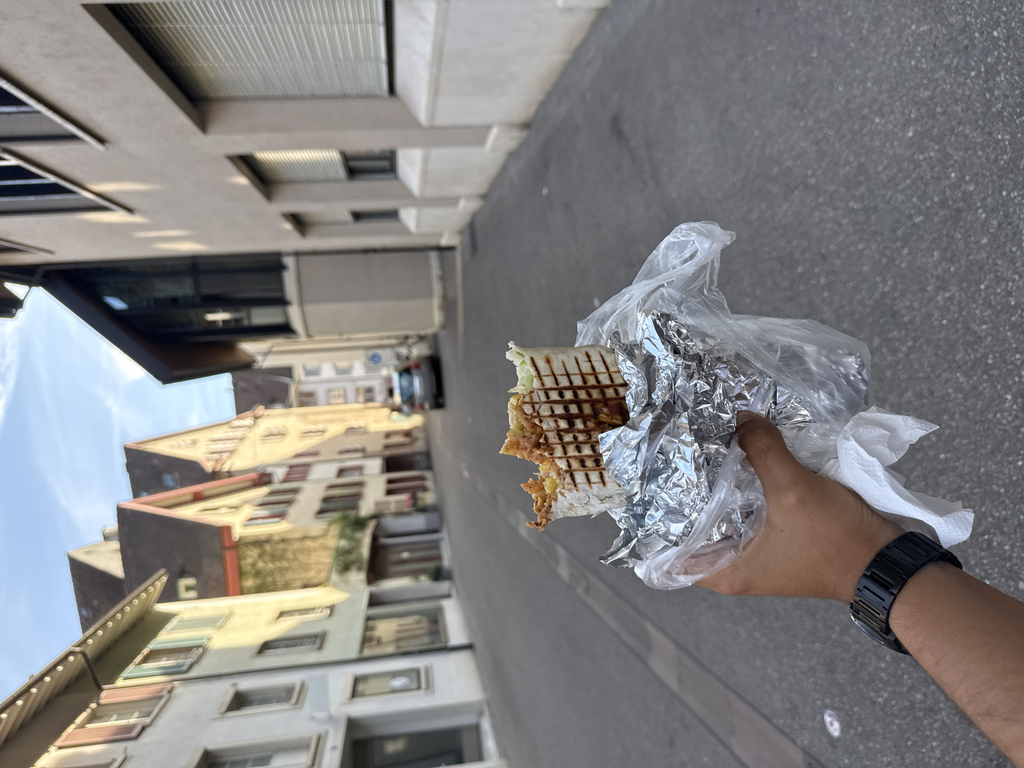 The image, correctly oriented, captures a person's hand holding a takeaway food item on a sunny street in Liestal, Switzerland. The central subject is a grilled wrap or sandwich, with visible char marks on the tortilla and some filling, including lettuce and other components, peeking out. It is partially encased in aluminum foil and a clear plastic bag. A black, possibly digital, wristwatch with a metal band is worn on the person's left wrist.
The scene unfolds in an outdoor urban setting, likely a street or alleyway. On the left, several traditional multi-story buildings line the street, showcasing varied facades in hues of yellow, brown, and off-white, characteristic of European architecture. Some feature pitched roofs and distinct window styles. On the right, the buildings appear more contemporary or commercial, with larger windows, some equipped with blinds, and a generally more uniform greyish-beige color palette. The street itself is paved with asphalt or a similar material.
The activity depicted is an individual holding a street food meal, suggesting they are about to eat or have just started. The bright, clear sky and distinct shadows indicate a sunny day, likely late morning or afternoon. No specific emotions are discernible, and no visible text can be read on the food packaging or buildings. The overall atmosphere is that of a pleasant, everyday moment in a Swiss town.