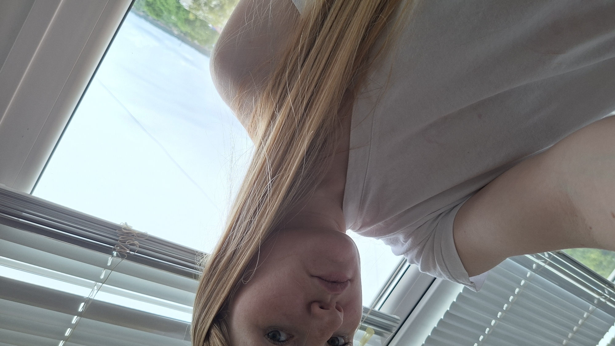 A young woman with long blonde hair is shown from the chest up, looking down and to the side. She is wearing a white t-shirt. The background consists of window blinds and a bright sky, with a hint of greenery visible outside. The image appears to be taken indoors, near a window.