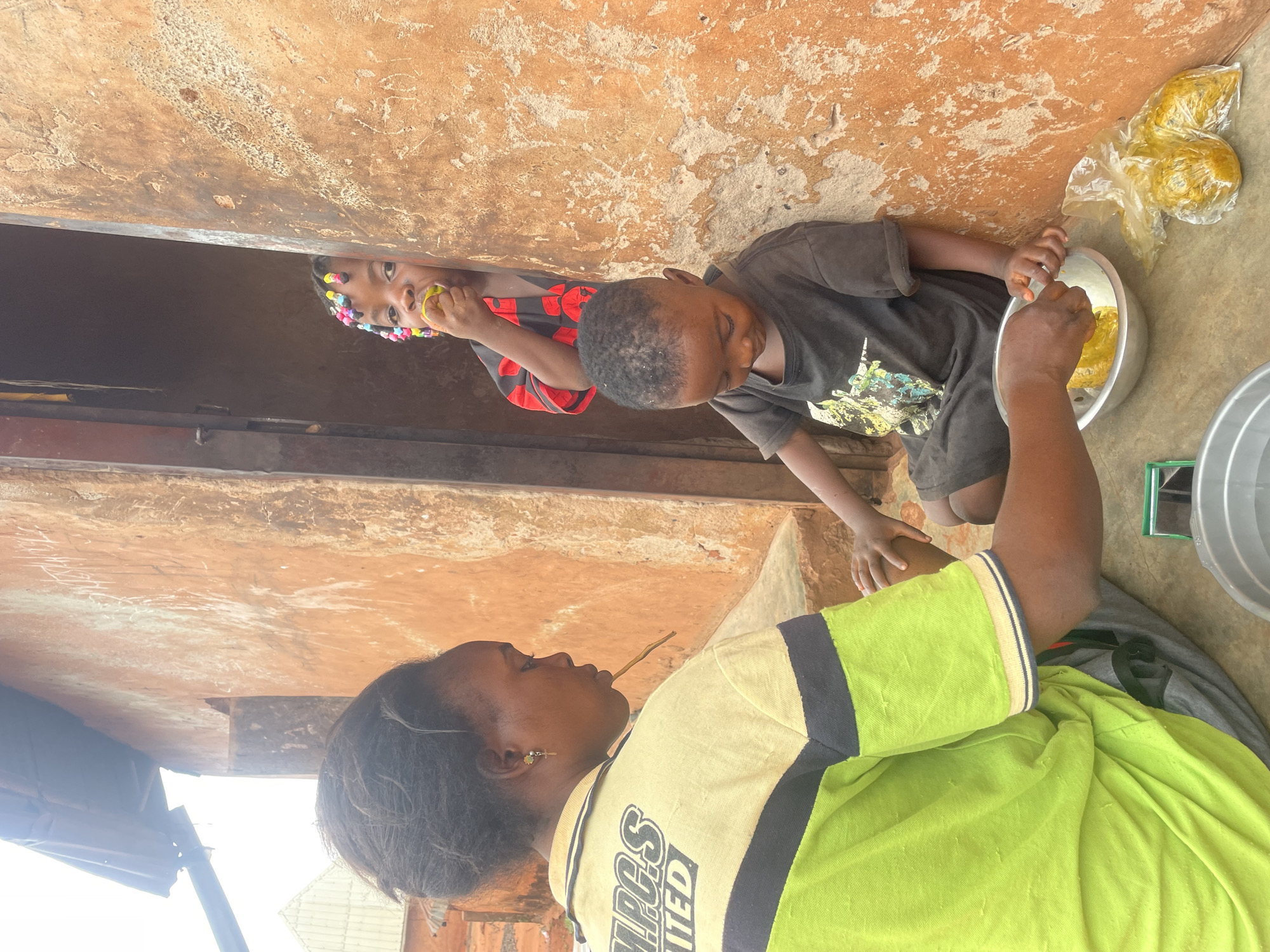 The image shows an outdoor scene in Ogbe, Nigeria. A woman wearing a green and yellow shirt with the inscription "NPGS UNITED" is seated, holding a thin, light-colored stick in her mouth. A young boy in a black t-shirt is beside her, bending over a silver bowl and handling yellow, wrapped food items. Another child is visible in the background, peeking through a window or doorway of a clay/mud building, also eating a yellow item. Beside the silver bowl are yellow food items in a plastic bag. A metal bowl sits to the right. The surroundings include a building constructed of orange/brown mud or clay with visible damage to the surface. The scene appears to capture a moment of food preparation or consumption in a somewhat rustic environment.