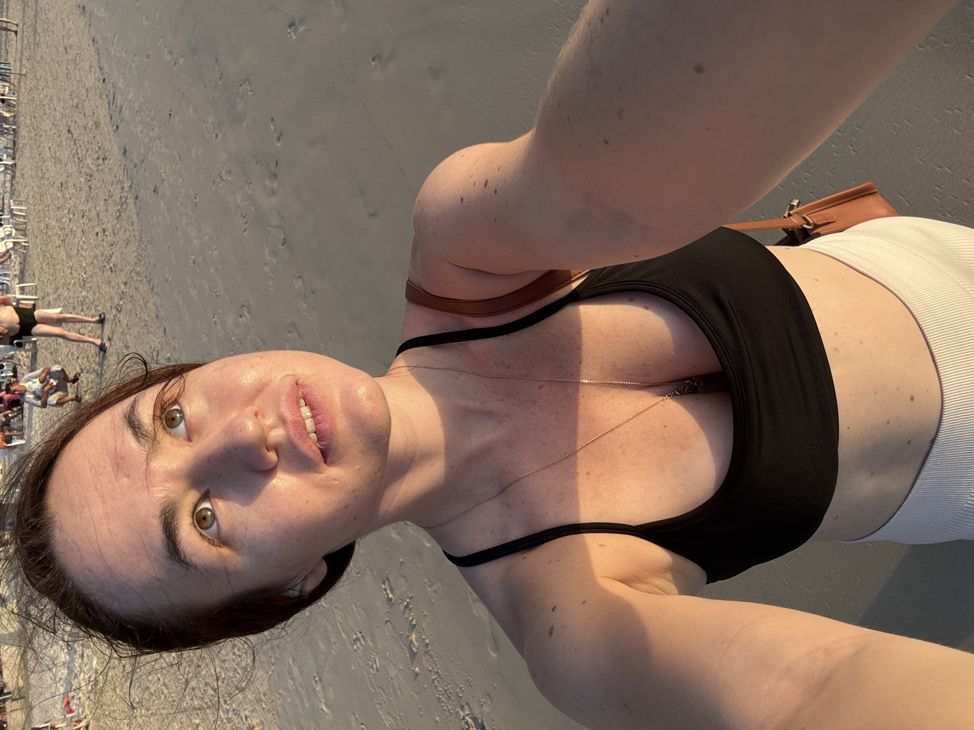 A woman is taking a selfie on a sandy beach in Patong, Thailand. The woman is wearing a black crop top and white pants. She has brown hair and brown eyes and is looking up and to the right. There are people in the background sitting on chairs and walking on the beach. The sun is setting, casting a warm glow on the scene.