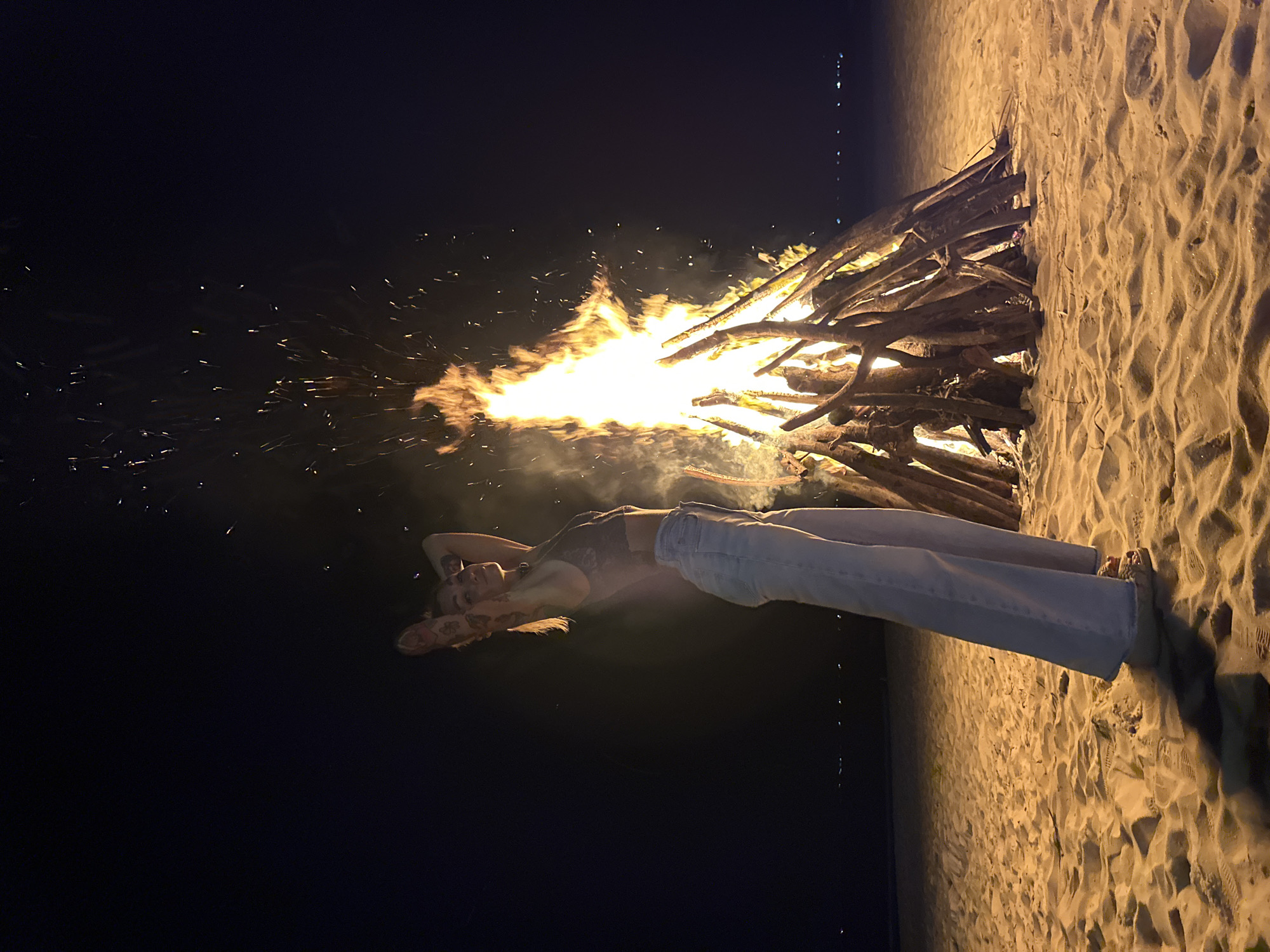 A young woman with blonde hair and tattoos on her arms stands on a sandy beach at night. She is wearing a dark lace crop top and light-wash, wide-leg jeans. Her arms are raised behind her head, and she is looking over her shoulder towards the viewer. To her right, a large bonfire burns, casting a warm glow on the scene and sending sparks into the dark sky. The beach is illuminated by the fire, and footprints are visible in the sand. In the background, the ocean is dark, with a faint line of lights suggesting a distant shoreline or city. The overall atmosphere is relaxed and serene. The location is identified as Phu Thuy Ward, Vietnam.