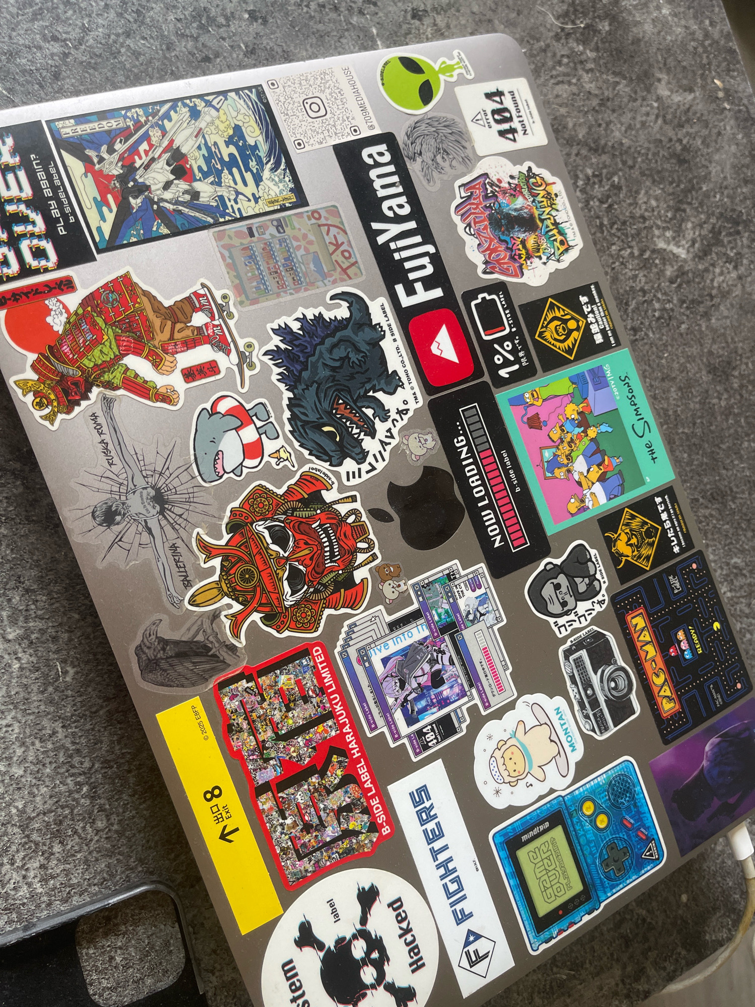 The image shows a close-up, overhead view of a laptop covered in numerous stickers. The stickers feature a variety of designs, including anime characters, Japanese text, popular culture references like "The Simpsons" and Pac-Man, a "404 Not Found" error, an alien, a gorilla, a shark, and a retro handheld gaming device. The laptop itself appears to be a MacBook, with the Apple logo partially visible. The surface the laptop rests on is a textured, grey, stone-like material, suggestive of an outdoor table or countertop. There is no indication of Paris, France, or any specific urban environment in the image. The scene is static, with no people or actions depicted.