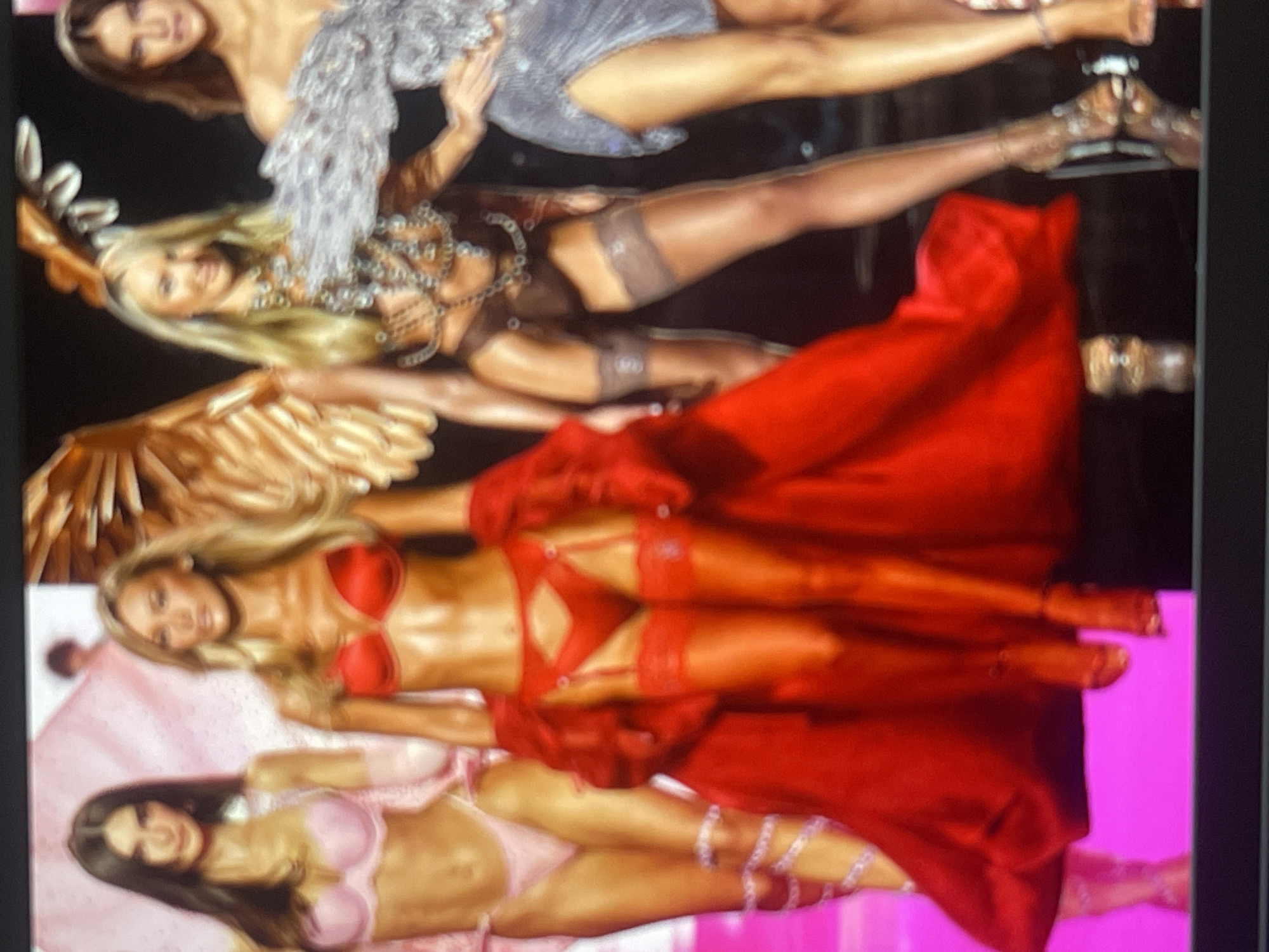 The media file displays a blurry, rotated image, likely a photograph of a screen, showcasing multiple female figures dressed in elaborate lingerie and costumes, indicative of a fashion show. The image is rotated 90 degrees counter-clockwise.
From top to bottom (when viewed upright):
The uppermost figure has brown hair and wears a sparkly, light grey or silver strapless garment. Below her is a blonde woman in a brown, embellished strapless top, accompanied by large, gold-colored "angel" wings. Both wear sheer stockings and high heels. The next figure is a blonde woman wearing a red lingerie set, complete with bra, garter belt, stockings, and a dramatic flowing red train or cape. The bottom figure has long dark hair and is dressed in a light pink bra and briefs, with sparkling adornments on her legs.
The setting appears to be a stage or runway with a dark background, possibly lit by spotlights, transitioning to a bright magenta or pink floor/backdrop in the lower section. The figures are posed dynamically, characteristic of fashion models presenting designs. The attire, particularly the "angel" wings and lingerie, strongly suggests an event similar to the Victoria's Secret Fashion Show. The overall impression is one of glamour and theatrical fashion. No visible text is present.
While the location context is provided as Poltava, Ukraine, there are no specific visual cues within the image itself to confirm this geographical detail. The image quality suggests it's a casual photograph of a digital display rather than an official event photo.