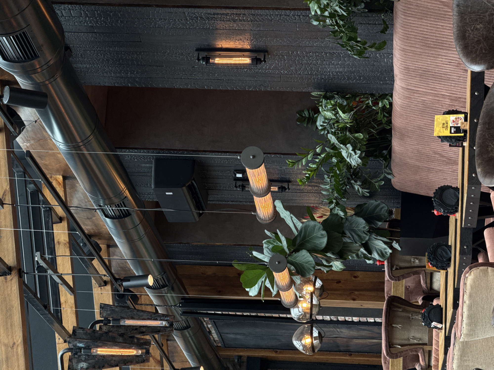 This image captures the interior of a restaurant in Izhevsk, Russia, characterized by an industrial-rustic design aesthetic. The scene presents an unoccupied dining area.
The upper section reveals exposed architectural elements, including dark metal beams, light-colored wooden planks, and a prominent dark metallic ventilation duct extending horizontally across the frame. Various lighting fixtures are suspended from these structures, including dark cylindrical track lights and decorative pendant lights. These pendants feature clear spherical glass globes housing exposed filament bulbs, and elongated cylindrical fixtures with ribbed casings, also containing visible filament bulbs. A large, black cubic speaker is mounted on the wall above the banquette.
The walls are clad in dark wood paneling, with the prominent right wall exhibiting a distinctly charred, textured finish. A wall-mounted light fixture with a caged, elongated filament bulb illuminates this dark wood.
The seating comprises both individual chairs and a banquette. Some chairs are upholstered in a light beige fabric with dark wooden frames, while a long banquette and adjacent chairs along the right wall are covered in a ribbed, reddish-brown fabric. Tables feature wooden tops supported by dark metal bases.
On the tables, black, fan-shaped napkin holders are visible, along with small red condiment shakers. A yellow menu card, displaying Cyrillic text that includes "ПЕТИТИ" (PETITI) and "БРЭКЕТ" (BRACKET), is present on one table.
Numerous large, leafy green plants, including a prominent fiddle-leaf fig, are strategically placed in dark square planters, integrating natural elements into the industrial setting. No individuals are present in the image.