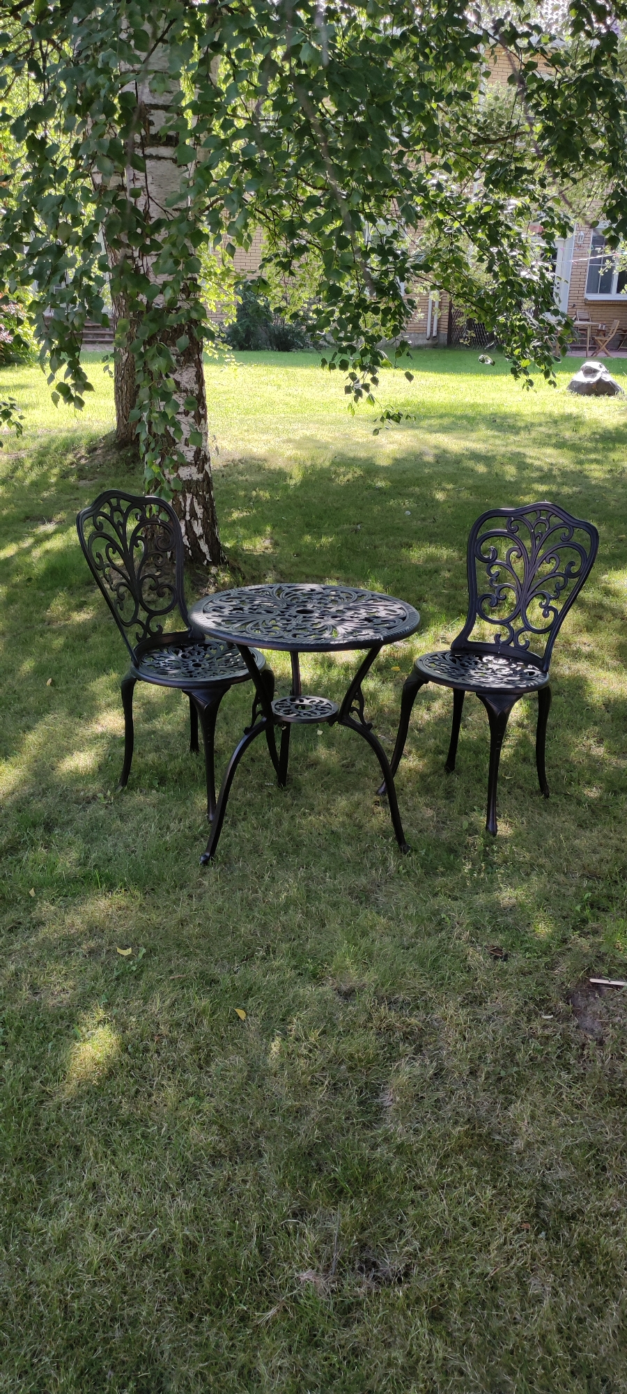 In Rosinka, Russia, a black wrought-iron table and two chairs are set on a lush green lawn beneath the dappled shade of a birch tree. The table is round with an ornate, floral-patterned surface and a lower shelf, supported by three curved legs. Each chair features a similarly decorative, high backrest with intricate scrolling designs and a seat matching the table's pattern, all supported by four slender legs. Sunlight filters through the tree's leaves, casting shadows across the grass. In the background, a light-colored brick house with visible windows and a darker-tiled roof is partially obscured by foliage. The overall scene depicts a tranquil outdoor seating area.