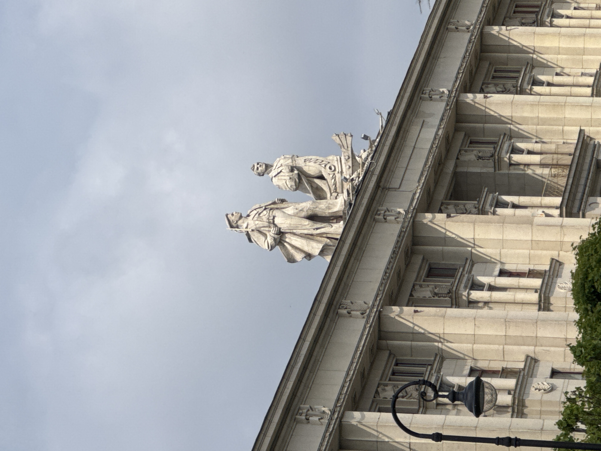 The image shows the facade of a building in Saint Petersburg, Russia, with two monumental sculptures of men on its rooftop. The building appears to be made of light-colored stone and features classical architectural elements such as columns and decorative carvings. The sculptures, also in light-colored stone, depict two male figures in military-style uniforms. One figure on the left is taller and appears to be holding a rifle or staff. The figure on the right is slightly shorter and stands with his hands clasped or held at his chest. They are positioned at an angle on the roof's edge. In the foreground, a black lamppost with a glass lantern is visible on the left side of the frame, and lush green foliage is present at the bottom. The sky is overcast with clouds. This building is likely the House of the Soviet of Leningrad Oblast (also known as the House of Soviets or the Soviet House) located at Ploshchad' Vosstaniya in Saint Petersburg, Russia, a prominent example of Stalinist architecture.