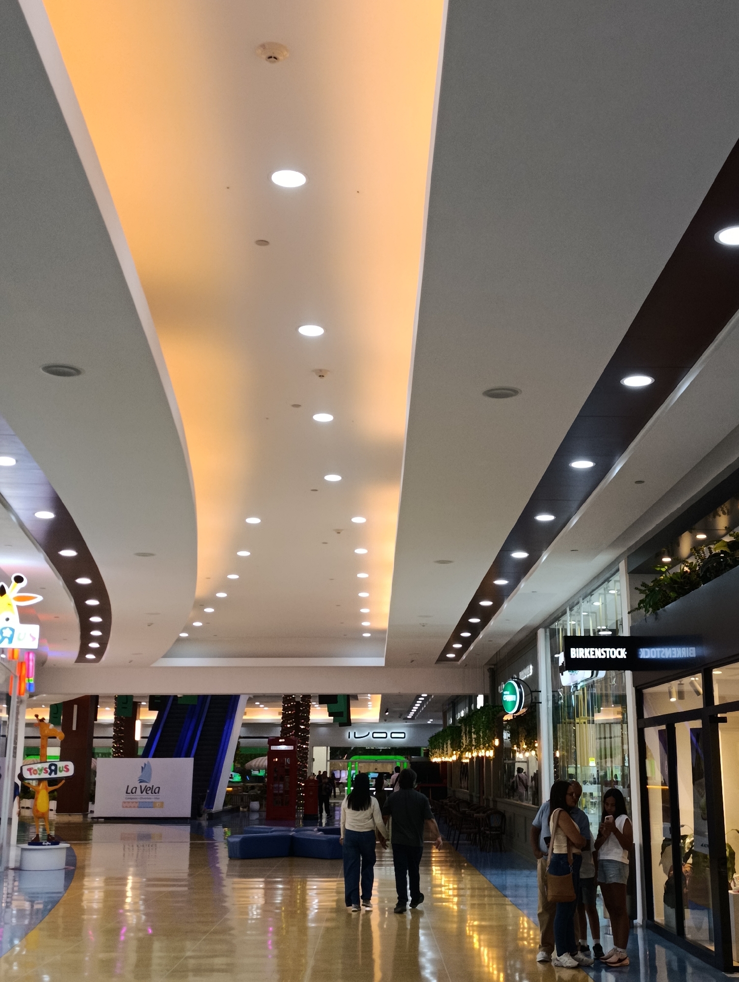 The image shows an indoor shopping mall in Porlamar, Venezuela. The mall has a high ceiling with multiple recessed lights arranged in rows. The ceiling has curved sections with different lighting designs. On the left side, a "Toys'R'Us" store is visible, along with an escalator. A "La Vela" sign is present. Several people are walking in the mall. On the right side, there's a "Birkenstock" store. A group of people are standing and talking near the store's entrance. The floor is glossy and reflects the overhead lighting.