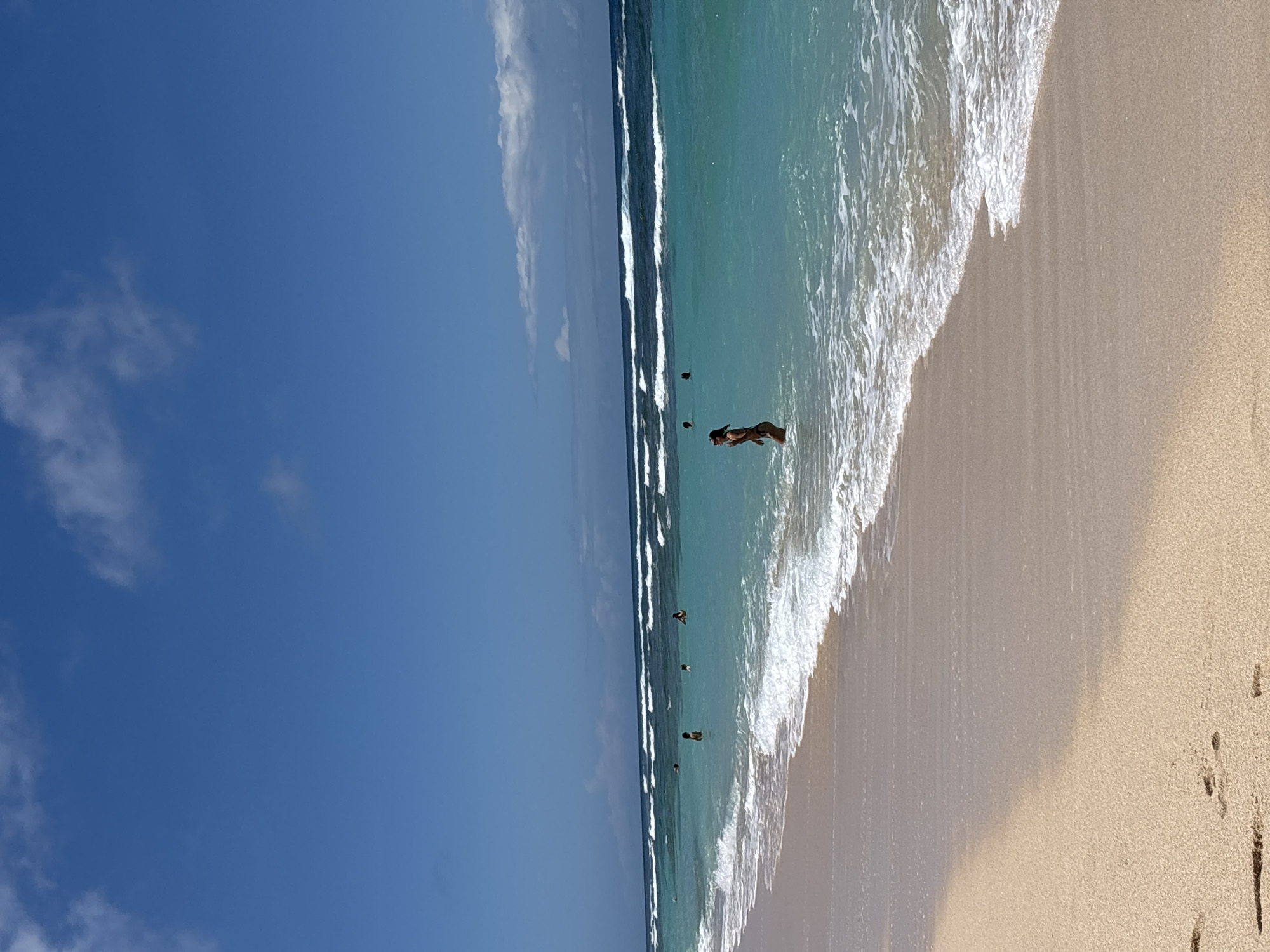 The image shows a beach scene in Ungasan, Indonesia. The sky is bright blue with some scattered white clouds. The ocean is a mix of turquoise and dark blue, with waves breaking along the shoreline. Several people are in the water, some closer to the shore and others further out. The beach is sandy, and footprints are visible in the sand near the edge of the water.