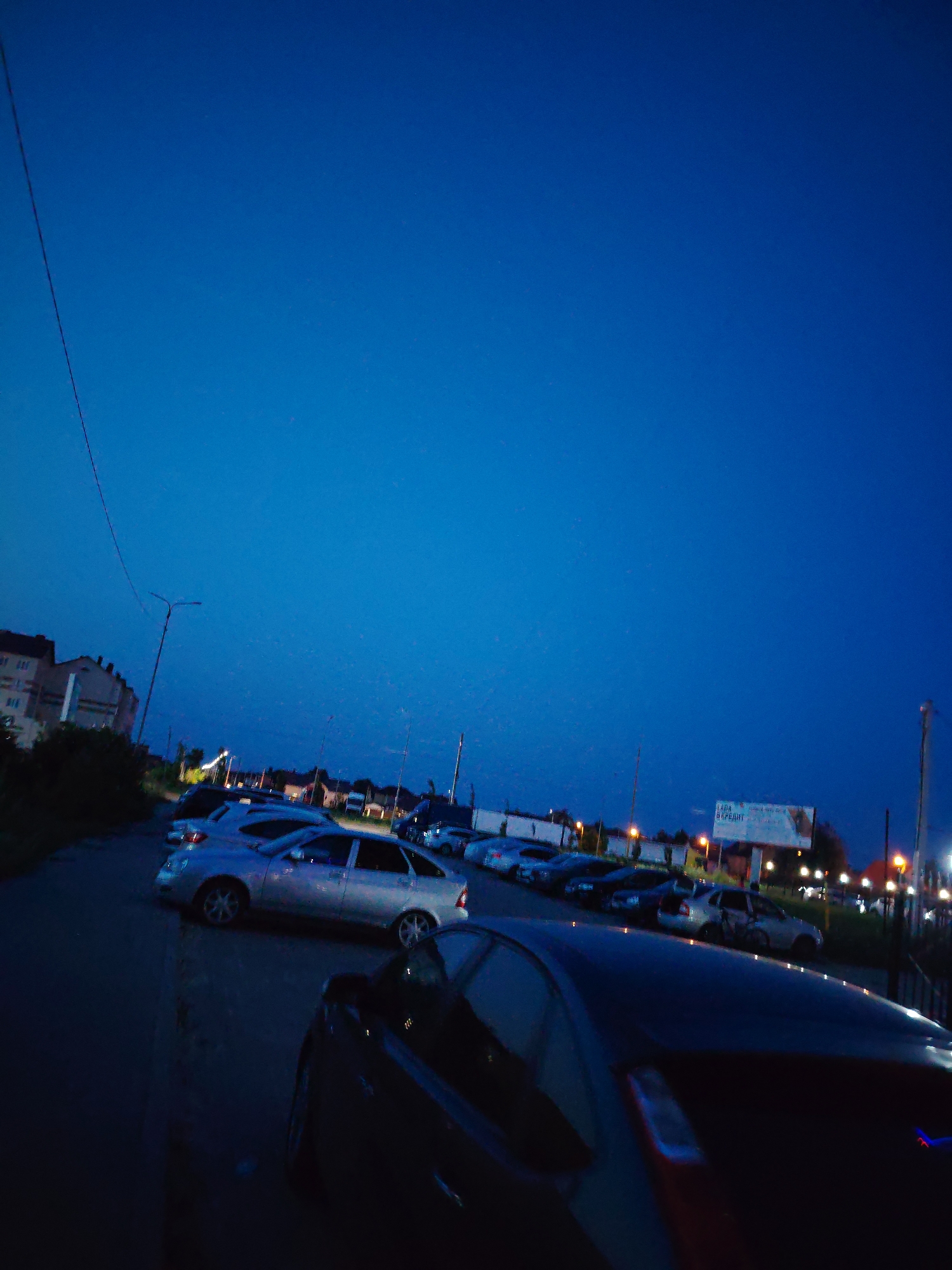 The image depicts a nighttime scene in a city, likely at dusk or early night. The sky is a deep, clear blue, indicative of twilight. A street lined with parked cars is visible, with a row of buildings on the left side of the frame. These buildings appear to be multi-story residential or commercial structures. Streetlights and lights from the buildings illuminate the scene. In the background, a billboard with Russian text is present, suggesting a commercial area. The overall atmosphere is calm and quiet, characteristic of a residential or suburban setting at night. It is impossible to determine the exact location of Крутые Выселки, Russia from the visual information provided.