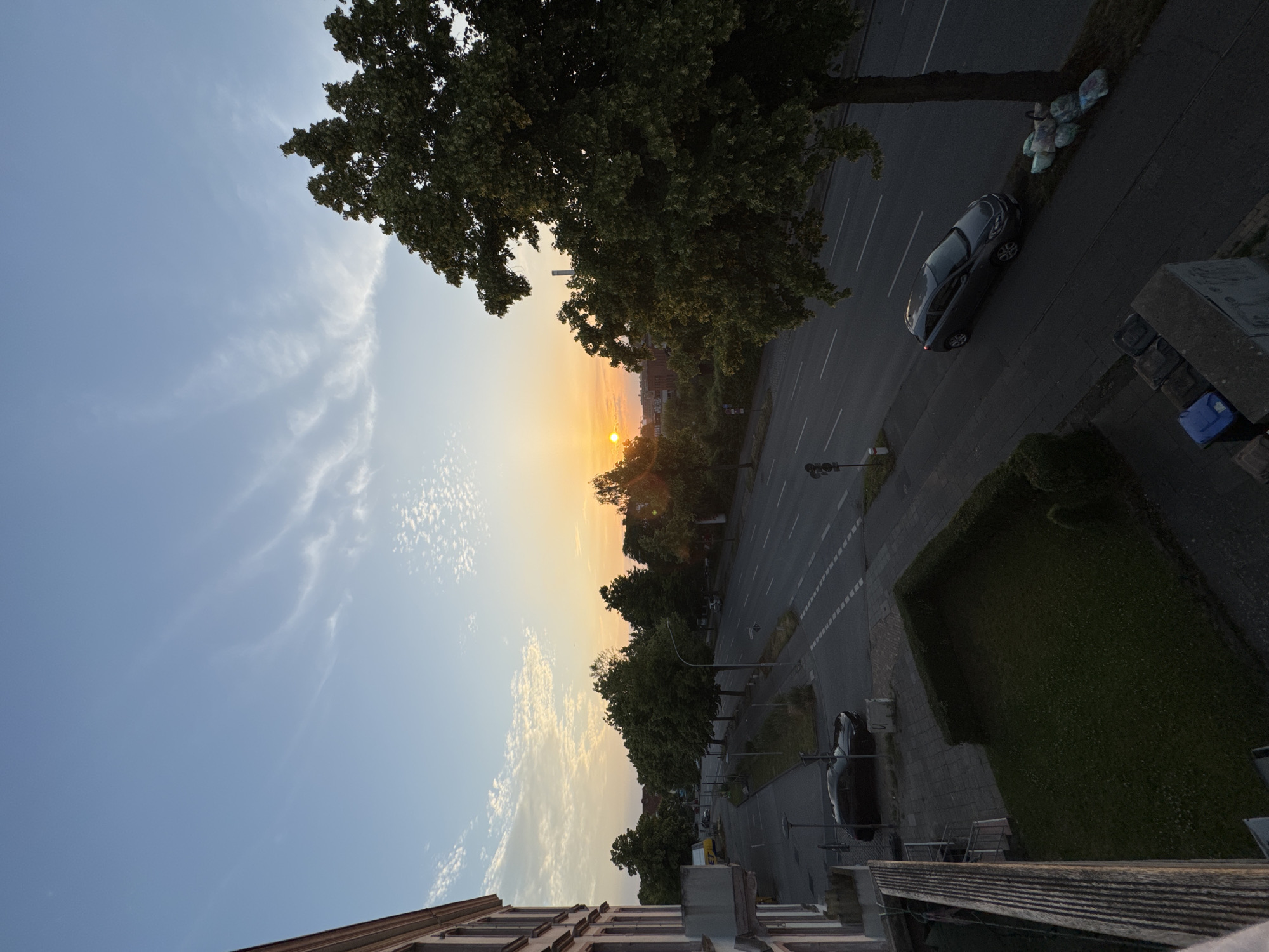 An elevated, eye-level, wide shot captures a street scene in Lübeck, Germany, at sunrise. The sky is a gradient of pale blue to soft orange, with wispy white clouds scattered across it. The sun, a bright orange orb, is positioned near the horizon, casting a warm glow over the scene.
In the foreground, a building's facade is visible on the left, its windows and balconies suggesting an urban residential area. A small patch of green lawn bordered by a manicured hedge is situated in the mid-ground. A dark-colored sedan is parked on the side of the road, and a row of garbage bags sits near the base of a large, leafy tree on the right.
The street itself is paved with asphalt and marked with white lane lines, indicating it is a multi-lane road. Traffic appears light, with only a few cars visible at a distance. Trees line the street, their green foliage contrasting with the urban structures and the warm tones of the sunrise. The overall impression is one of a peaceful, early morning in a city setting.