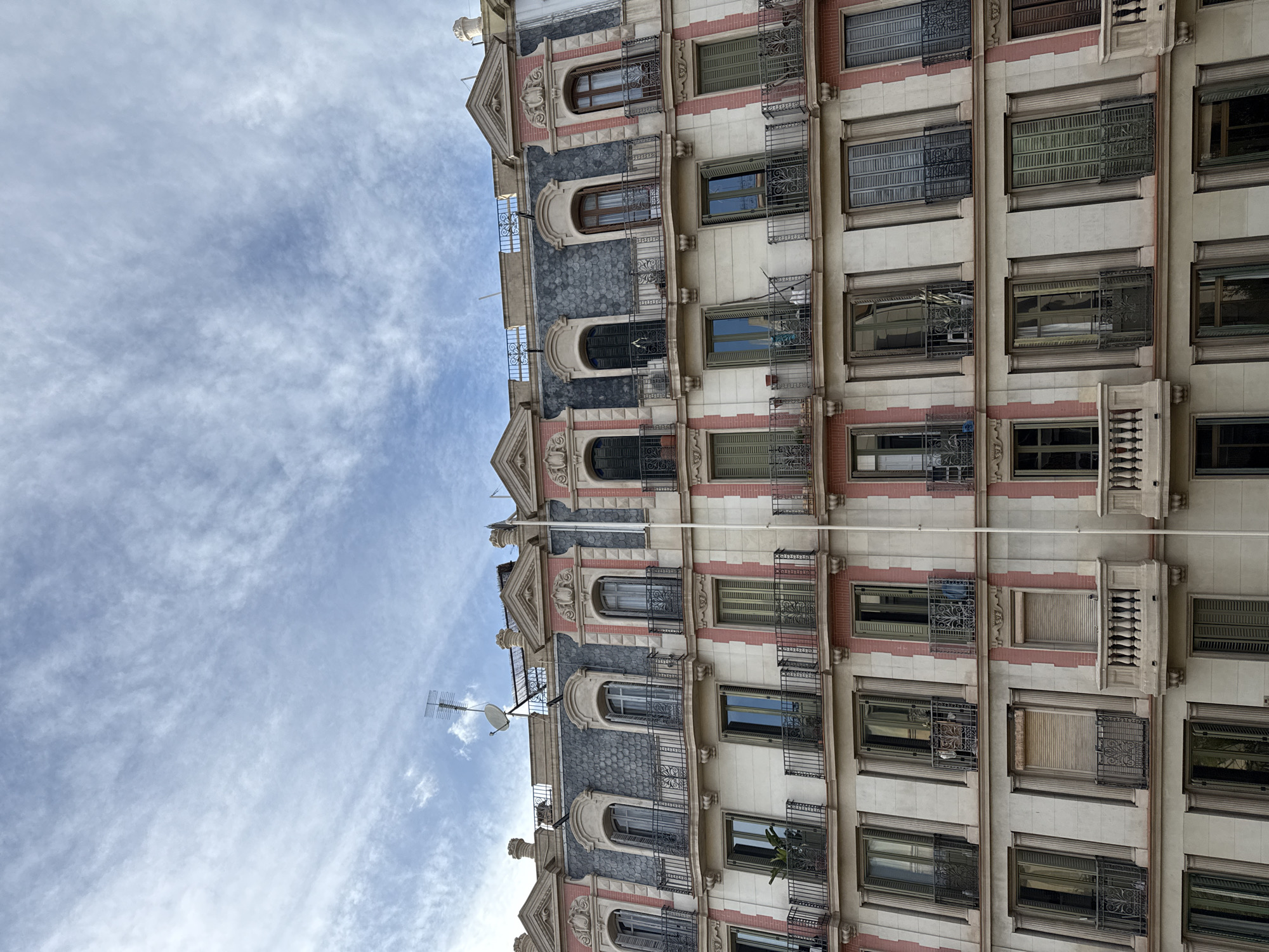 The image displays the upper seven stories of a classical multi-unit residential or commercial building facade, characteristic of Barcelona's Eixample district, Spain. The structure is constructed primarily from light-colored stone or stucco, featuring vertical sections of pale pink brickwork and decorative elements.
The uppermost two stories are integrated into a dark grey mansard roof, possibly slate-tiled. This level features a series of arched windows with dark frames, several of which are open, leading to small balconies with black wrought-iron balustrades. Ornate pediments adorn the tops of these windows. On the rooftop, a silver television antenna and a small satellite dish are mounted.
Below the mansard, the next two levels present rectangular windows with dark frames and predominantly dark green louvered shutters, many in an open position. These windows also lead to balconies with black wrought-iron balustrades. A bicycle is visible stored on one of these balconies on the second level from the top.
The lower three visible stories continue the pattern of rectangular windows, some with light frames, and a mix of dark green and brown louvered shutters, with varying states of open and closed. Some windows have closed roller shutters. Most balconies on these levels feature black wrought-iron balustrades, but one prominent balcony on the third level from the bottom (left side) is distinguished by a cream-colored stone balustrade. A single white vertical pole extends down the facade slightly to the right of the center. Potted plants are visible on several balconies.
The sky above the building is light blue with scattered, thin cirrus clouds. No people are visible within the frame.