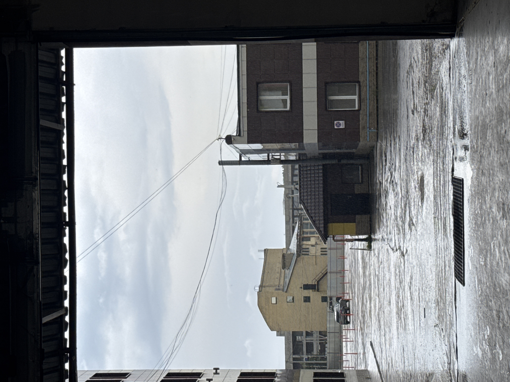 The image displays an exterior scene from inside a dark, framed opening, likely a garage door, looking out onto a wet, paved urban environment. The ground in the immediate foreground is wet concrete with a rectangular drainage grate. Beyond the frame, a large, puddled asphalt or concrete lot extends, marked by numerous red and white bollards. Two parked vehicles, a dark SUV and a lighter-colored truck, are visible near a corrugated metal fence in the midground. The sky is overcast and grey, with several utility lines traversing it.
Architectural elements dominate the background. On the far left, a multi-story, light-colored building with dark window openings appears to be under construction or renovation. Central to the frame is a prominent, light-tan brick building featuring an irregular roofline, multiple small windows, and an external metal staircase on its right facade. Behind this, skeletal structures indicate further construction. To the right, a dark brown, multi-story building with white horizontal banding displays two windows and a blue 'No Parking' sign. A utility pole with associated wiring stands beside it. Adjacent to this building and closer to the foreground is a smaller, single-story structure with a dark tiled roof and a section of yellow wall. The overall scene suggests a recent rainfall event in an industrial or commercial district of Perm, Russia. No people are visible.