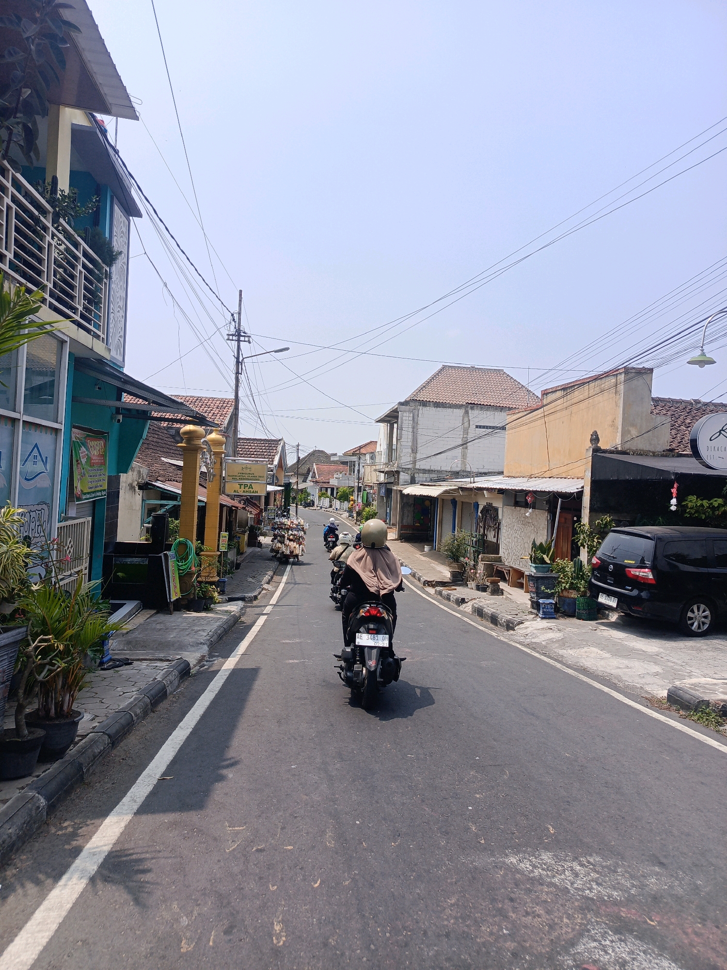 The image depicts a street scene in Kebonagung, Indonesia. The perspective is from behind a person riding a motorcycle on a paved road. The road has a white line dividing the lanes. On the left side of the road, there are buildings with multi-level structures, balconies with railings, and plants in pots. Some of these buildings appear to be shops or residences, with signage and awnings visible. On the right side of the road, there are more buildings, also with awnings and some with plants. A black car is parked on the right side of the road. Several other motorcycles are visible further down the road, also with riders. The sky is clear and pale blue. Utility poles with numerous wires are present above the buildings and road. The overall impression is a moderately busy urban street on a clear day.