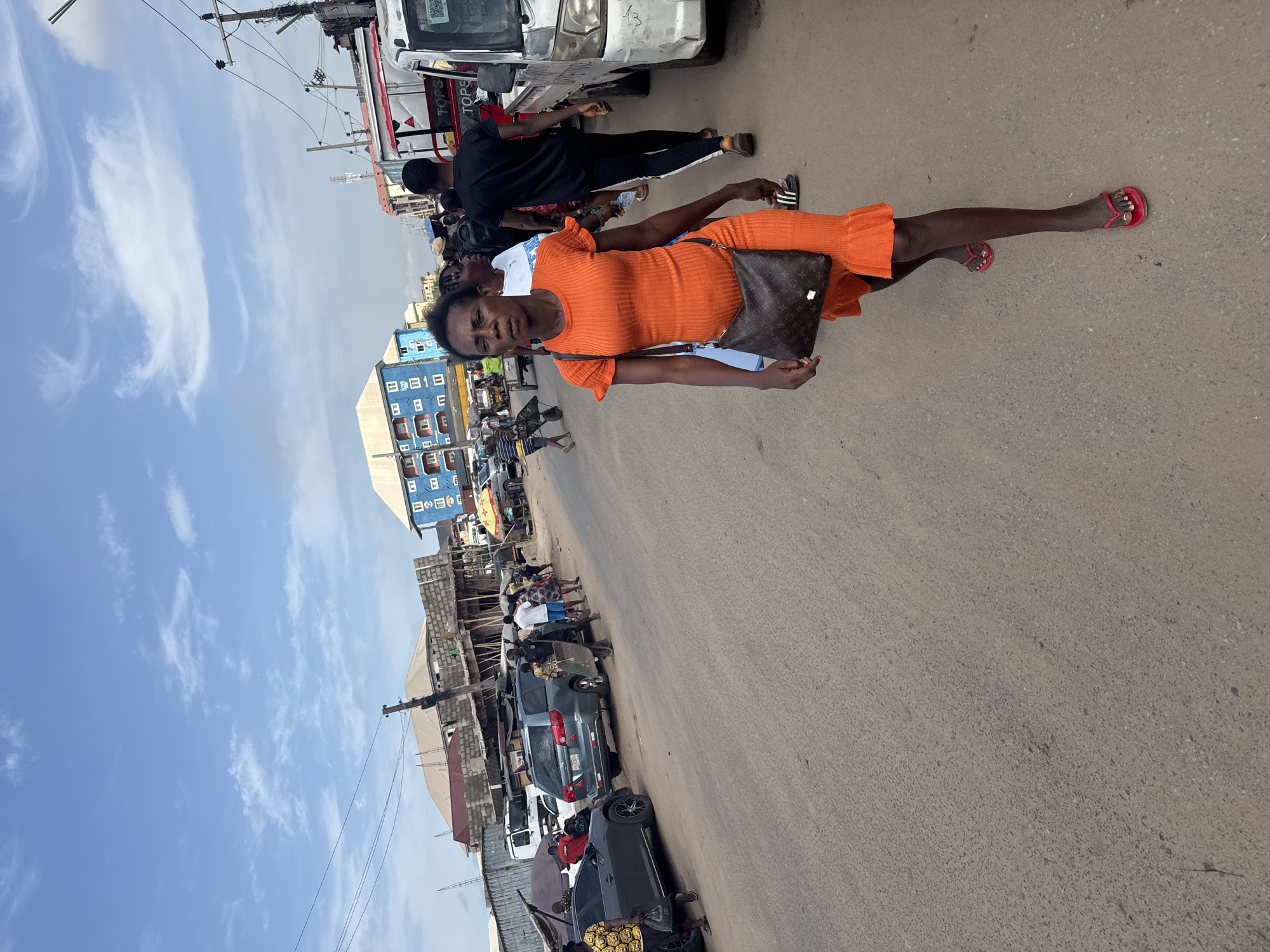 The photo is taken at street level in Onitsha, Nigeria. A woman in an orange dress and red sandals stands on the street. She carries a brown patterned purse. Behind her, a man in black clothing walks next to a white vehicle. Further back, a building with blue and white facade is visible. Cars are parked along the street, and other people are moving around. The sky is blue with scattered clouds. The road surface appears to be asphalt.