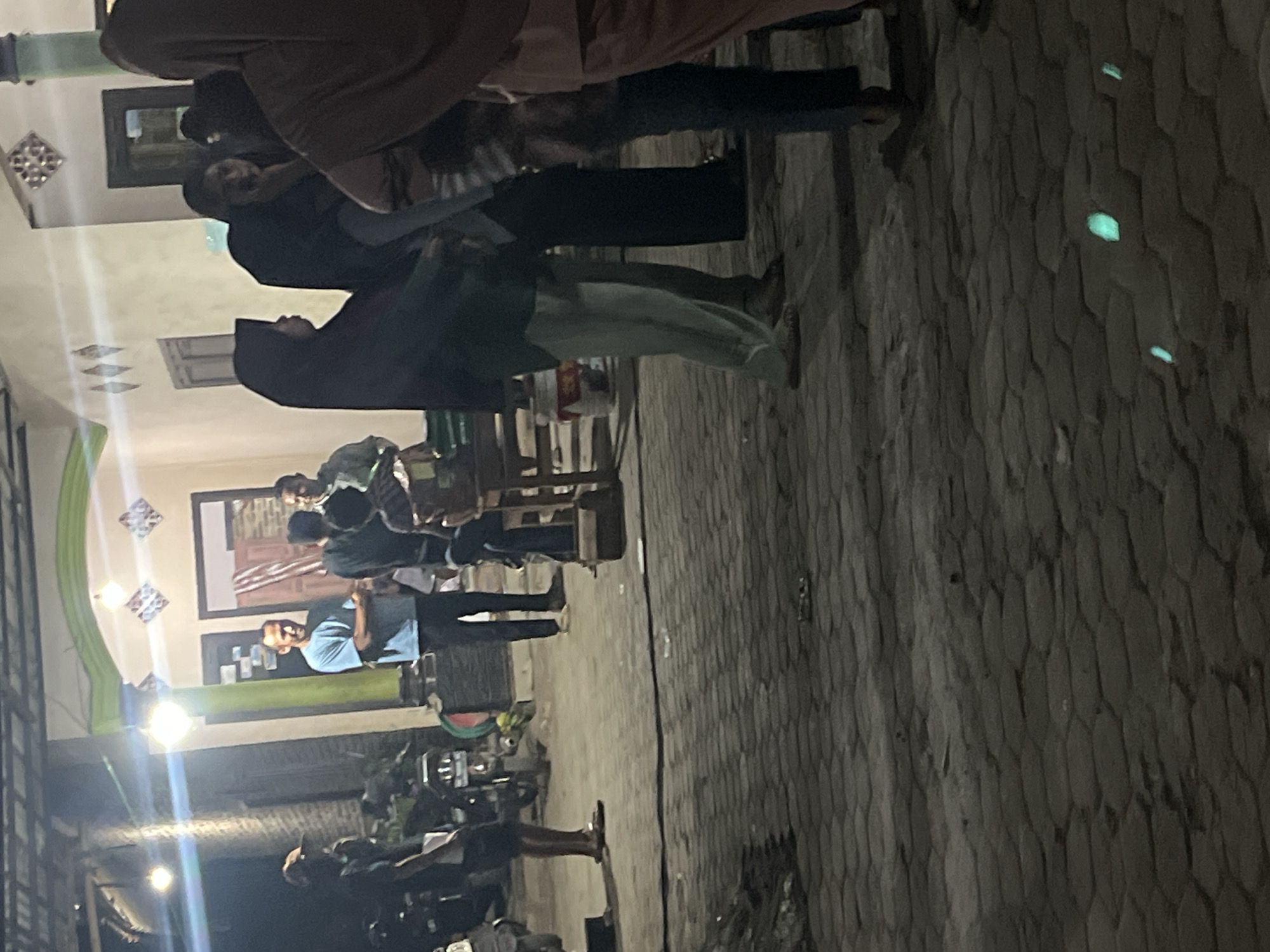 A group of people are gathered on a paved outdoor area in front of a building at night. Several individuals are standing and talking, while others are seated on chairs. A motorcycle is parked to the left of the frame. The building appears to be a residence or commercial establishment, with lights illuminating the facade. The ground is paved with interlocking stones. The lighting suggests an evening or nighttime scene, possibly in Kebumen, Indonesia, given the architectural style and commonality of such scenes in the region.