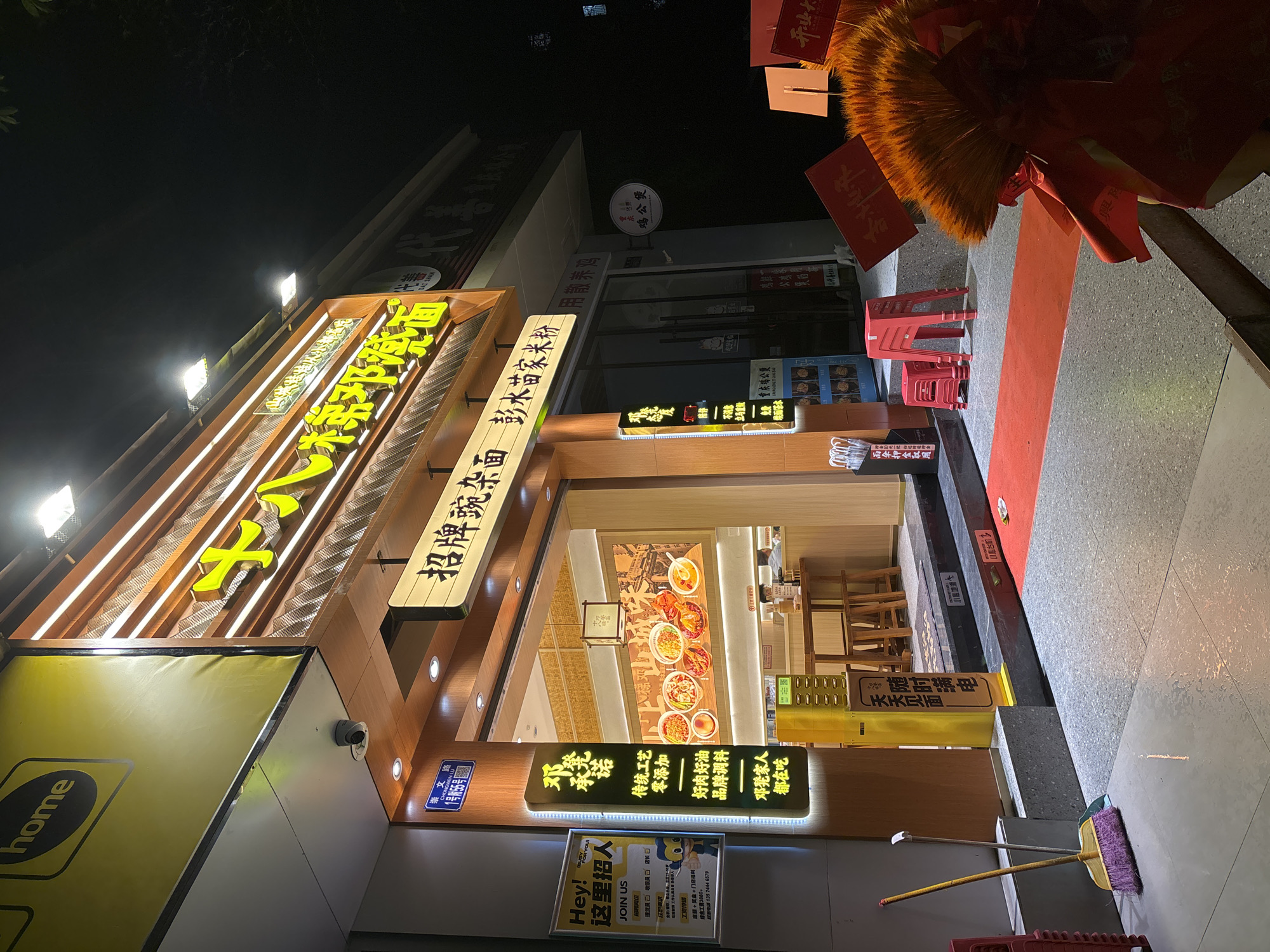 This is a nighttime view of a brightly lit restaurant front. The restaurant appears to be located in the Nan'an District, China, as a sign indicates "CHONG 文路1号附55号", which translates to "Chong Wen Road, No. 1, attached 55" in Chinese. The restaurant has prominent signs in Chinese characters, some illuminated with neon lights. The storefront is decorated with traditional red decorations and a red carpet leads into the restaurant's entrance. Several red plastic chairs are stacked nearby. Two brooms lean against the wall near the entrance. A sign advertising job openings at the restaurant is posted outside. The restaurant's interior is visible through the open entrance, revealing tables and chairs.