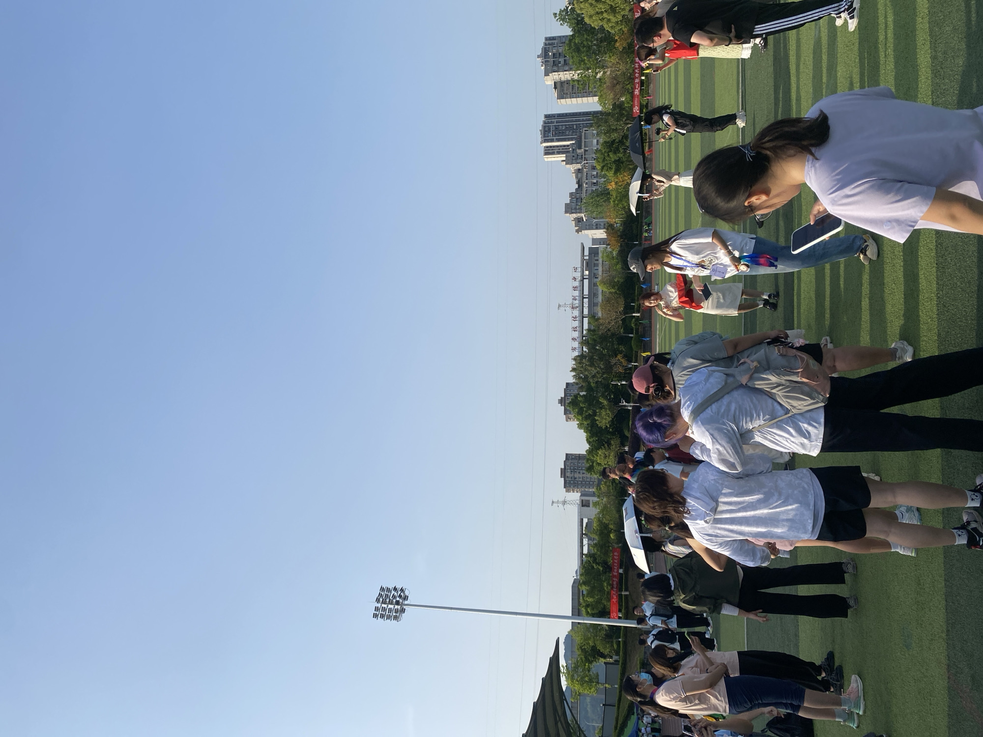 The image shows a group of people standing on a green field, possibly a sports field or park area. In the background, there are buildings and trees, indicative of an urban environment. A tall light pole with multiple lamps is visible on the left side of the image. The location is in Chongqing High-tech Zone, China. People appear to be standing, possibly observing an event or participating in an activity.