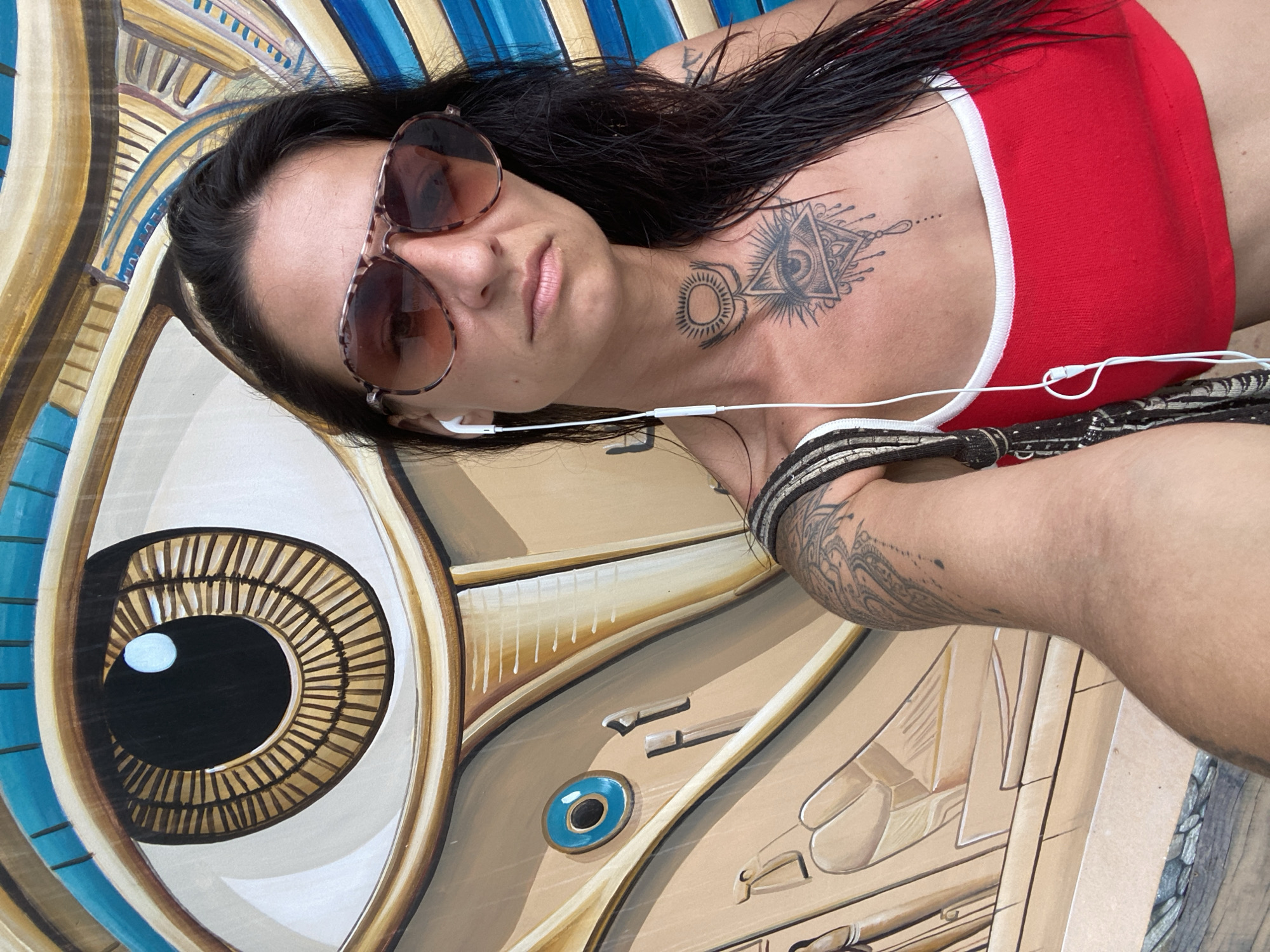 A woman with long, dark, wet hair and a red top is taking a selfie in front of a large, colorful mural. She is wearing aviator sunglasses with a tortoiseshell pattern and white earbuds. There are several tattoos visible on her chest and arm, including an eye within a triangle on her chest. The mural behind her features a stylized, large eye with concentric circles and Egyptian motifs, rendered in warm earthy tones and bright blues. The overall impression is of a public art installation.