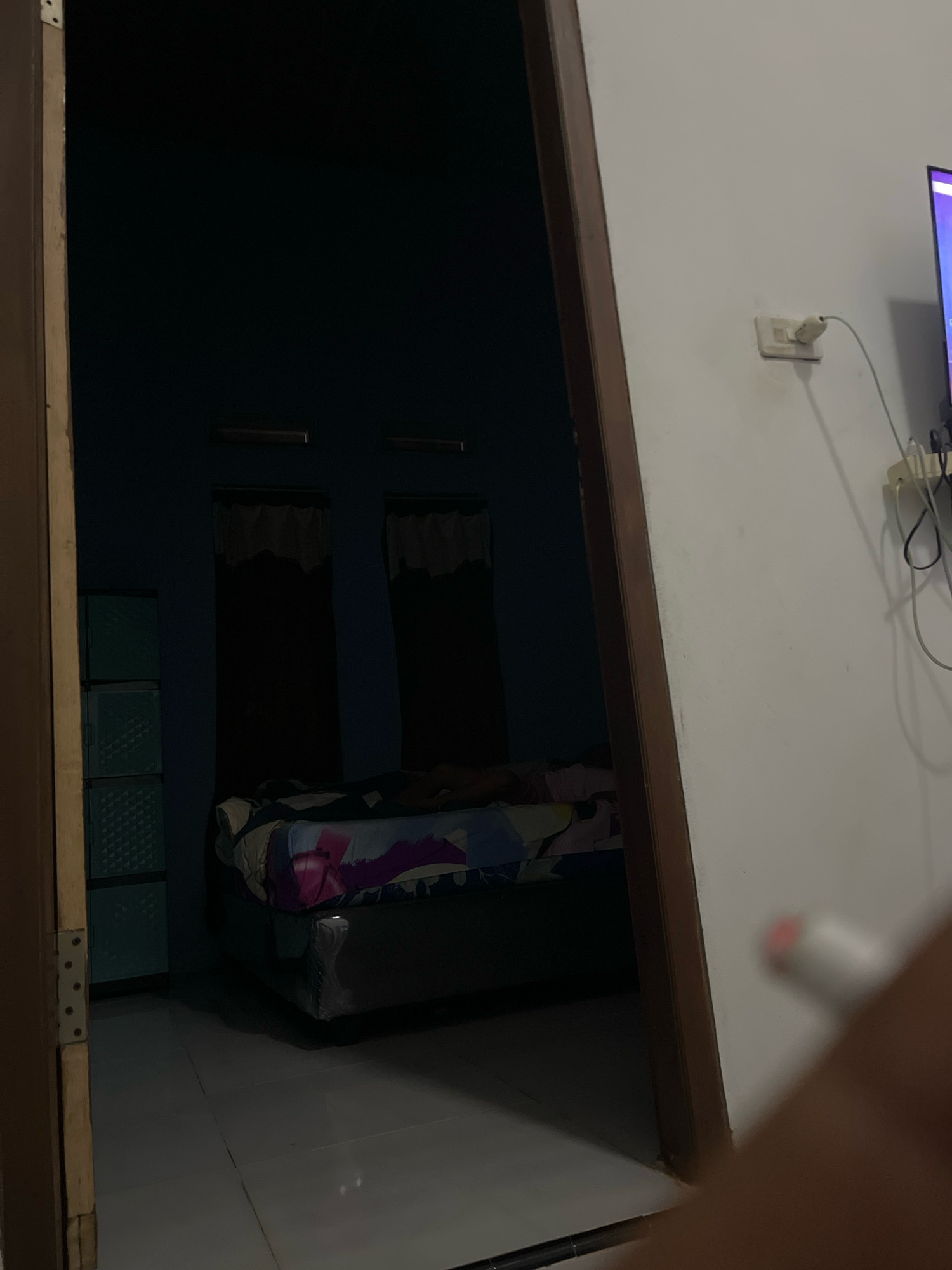 The image shows the interior of a dimly lit bedroom, viewed from a doorway. On the right side of the frame, a white wall is visible with a light switch and a power outlet with a cord plugged into it. Further to the right, a portion of a television screen displaying a bright purple color is visible. On the left side of the frame, within the room, a light blue plastic cabinet with three drawers is present. In the center of the room, a bed with a colorful patterned comforter is visible. The floor is tiled. A person's hand holding a white object with a pink tip is partially in the foreground on the right side of the image. The scene appears to be in a residential setting in Kebumen, Indonesia.