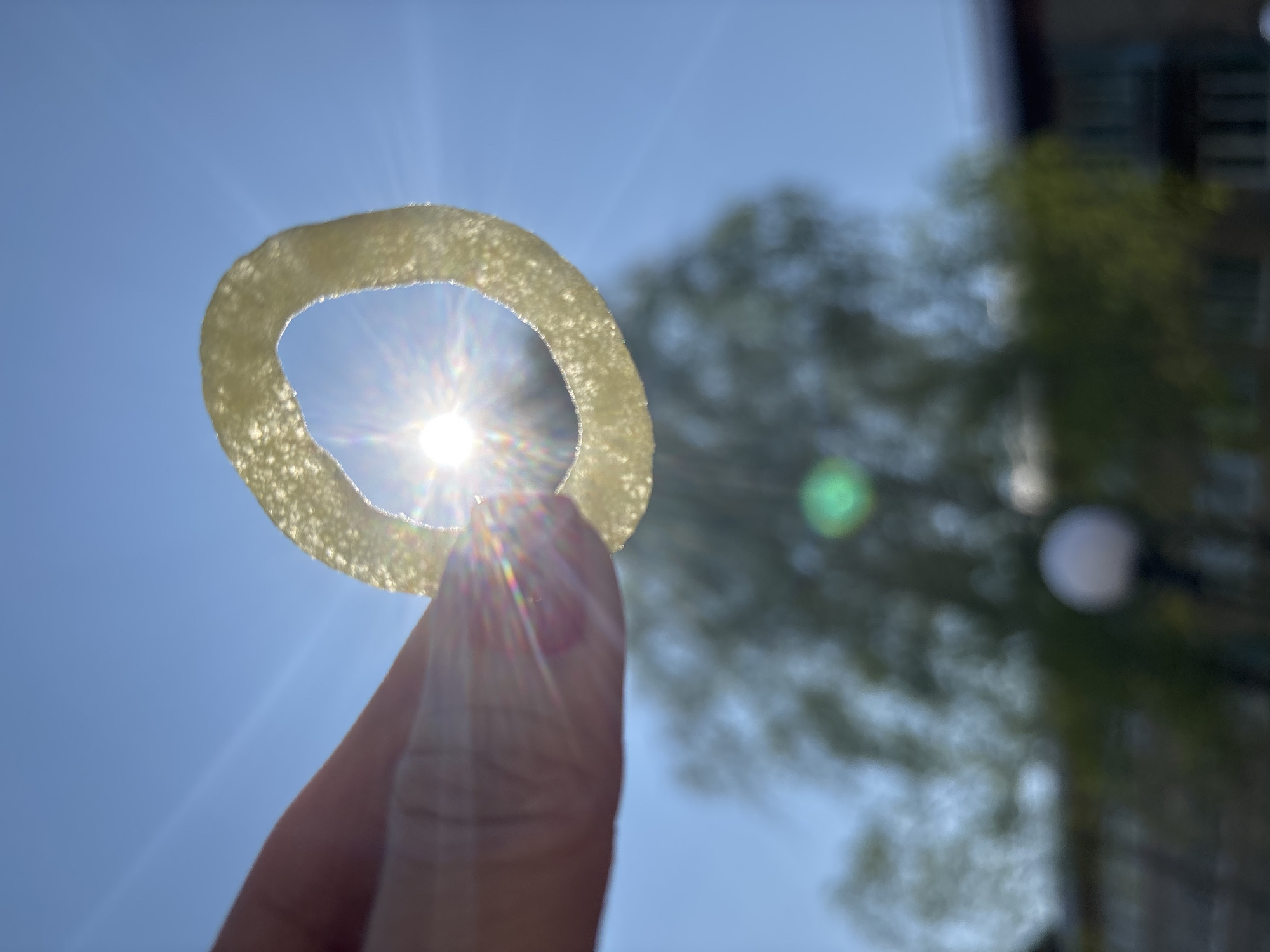 A hand holds up a translucent, rough-textured ring against a bright blue sky, with the sun shining through its center, creating a starburst effect. In the blurred background, there is greenery that appears to be a tree and portions of a building. There is no confirmation that the photo was taken in the city of Chaikovsky, Russia, and there is no indication of actions or interactions. The background environment is blurred and not distinctly identifiable.