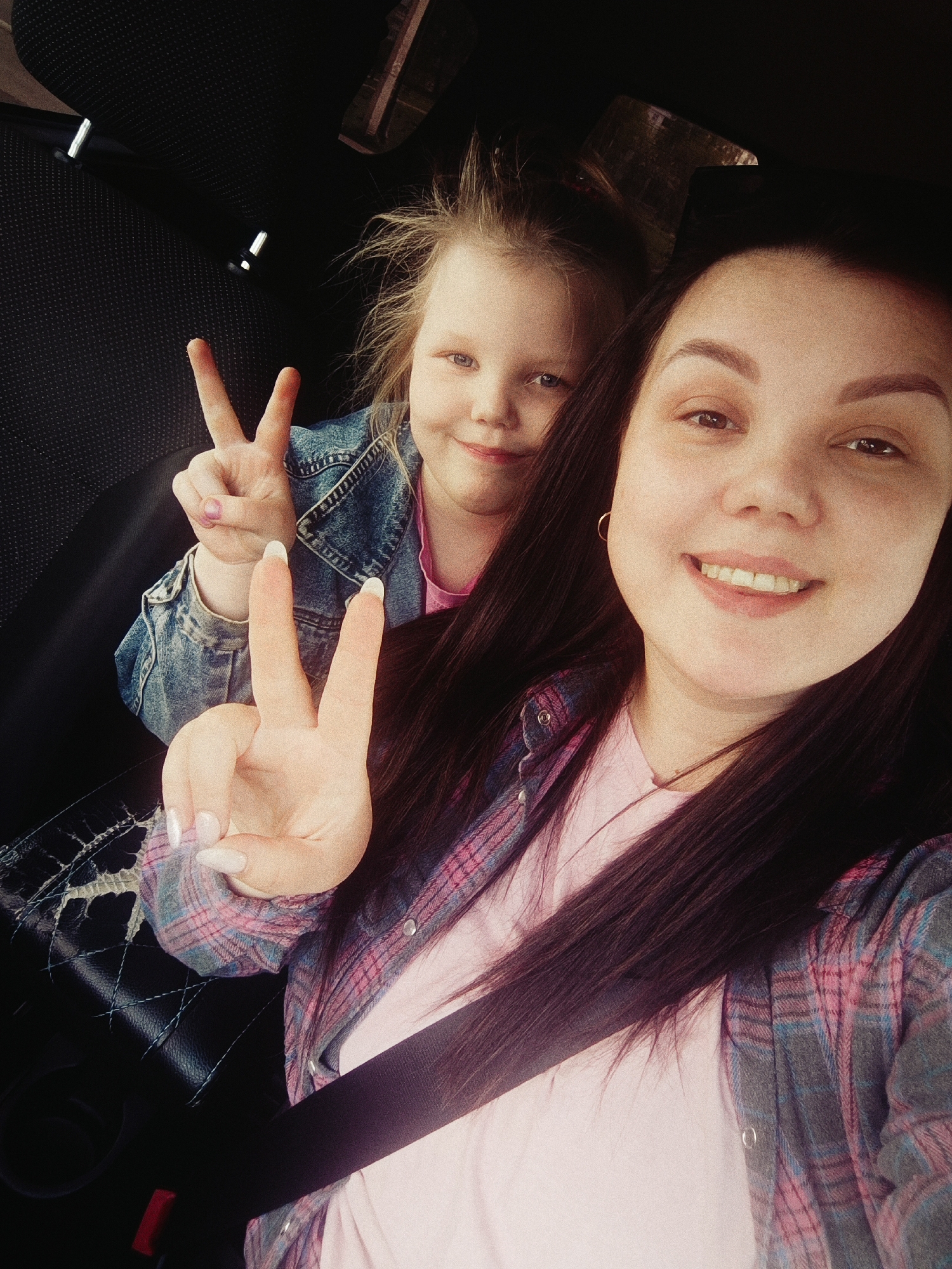 A woman and a young girl are posing for a selfie inside a car. The woman, in the foreground, is wearing a pink t-shirt and a gray and pink plaid flannel shirt. She has long, dark brown hair, and she is smiling. A seatbelt is visible across her body. Her right hand is raised, making a peace sign. The young girl is seated behind the woman, also making a peace sign. She is wearing a pink t-shirt and a blue denim jacket. The interior of the car appears dark, with black upholstery visible. The location is Chaikovsky, Russia.