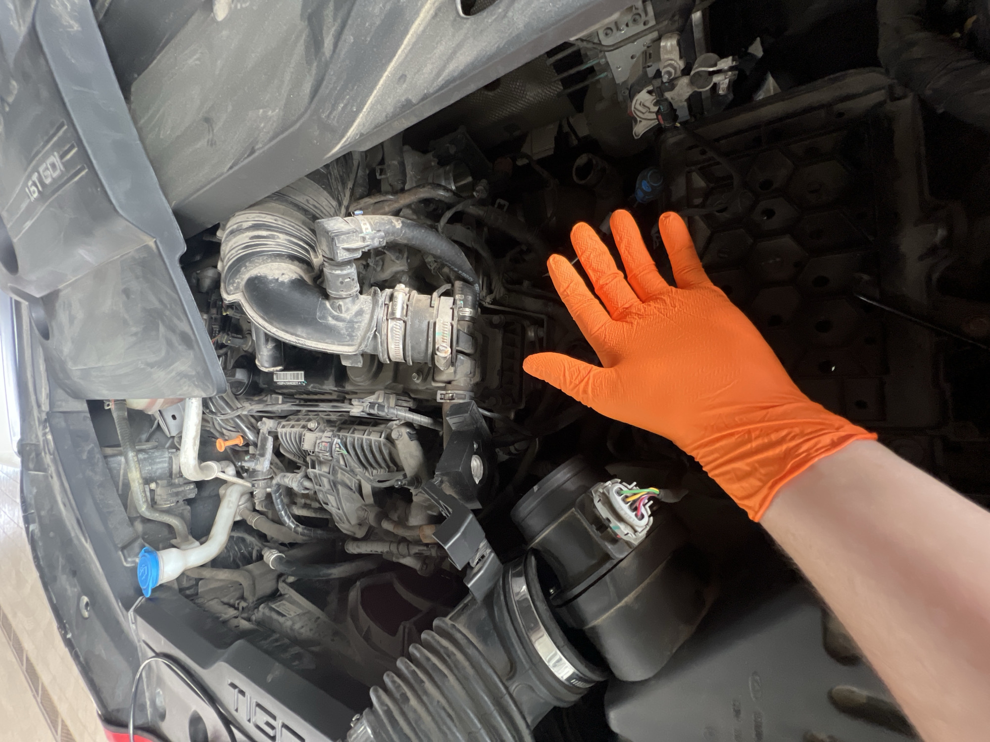 An overhead view captures the open engine bay of a Chery Tiggo vehicle in Izhevsk, Russia, likely during maintenance or inspection. A person's right forearm, clad in a bright orange protective glove, extends into the lower right portion of the engine compartment, with the palm facing upwards. The engine bay exhibits a moderate accumulation of dust and grime on various components.
Key visible components include a white plastic windshield washer fluid reservoir with a blue cap in the upper left, an orange-capped dipstick handle near the engine block, and a prominent black corrugated air intake hose connected to a sensor with multicolored wiring (green, yellow, white, red, black) in the lower left. A large black plastic engine cover, partially removed or repositioned, is visible in the upper right, bearing the embossed text "1.6T GDI," indicating a 1.6-liter Turbo Gasoline Direct Injection engine. Another plastic cover on the far left bears the partial text "TIG," further identifying the vehicle as a Chery Tiggo. Numerous other hoses, wires, and metallic components are visible throughout the cluttered engine space. A hexagonal-patterned dark plastic cover, possibly for a battery or fuse box, is present in the lower right quadrant of the image.