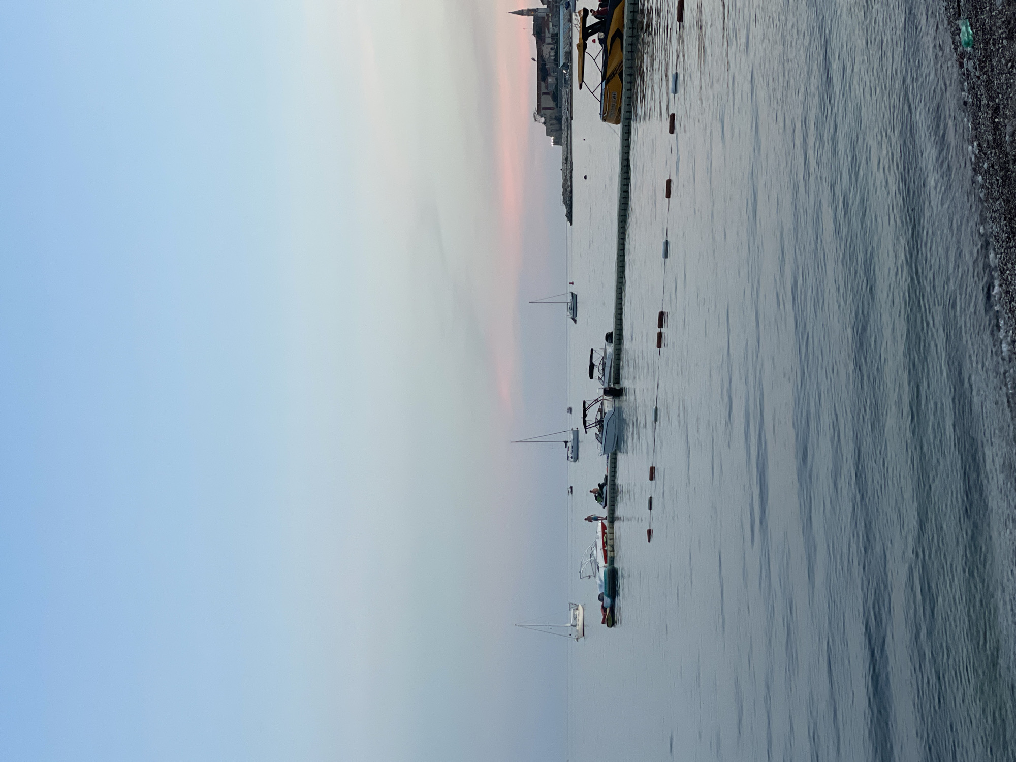 The image shows a calm body of water with several boats docked. A floating walkway with buoys extends into the water. In the background, a town with a church spire is visible on a peninsula. The sky is a soft gradient of blue and pink, indicating either dawn or dusk. There are a few people on the dock and on one of the boats. The foreground shows a pebble beach.