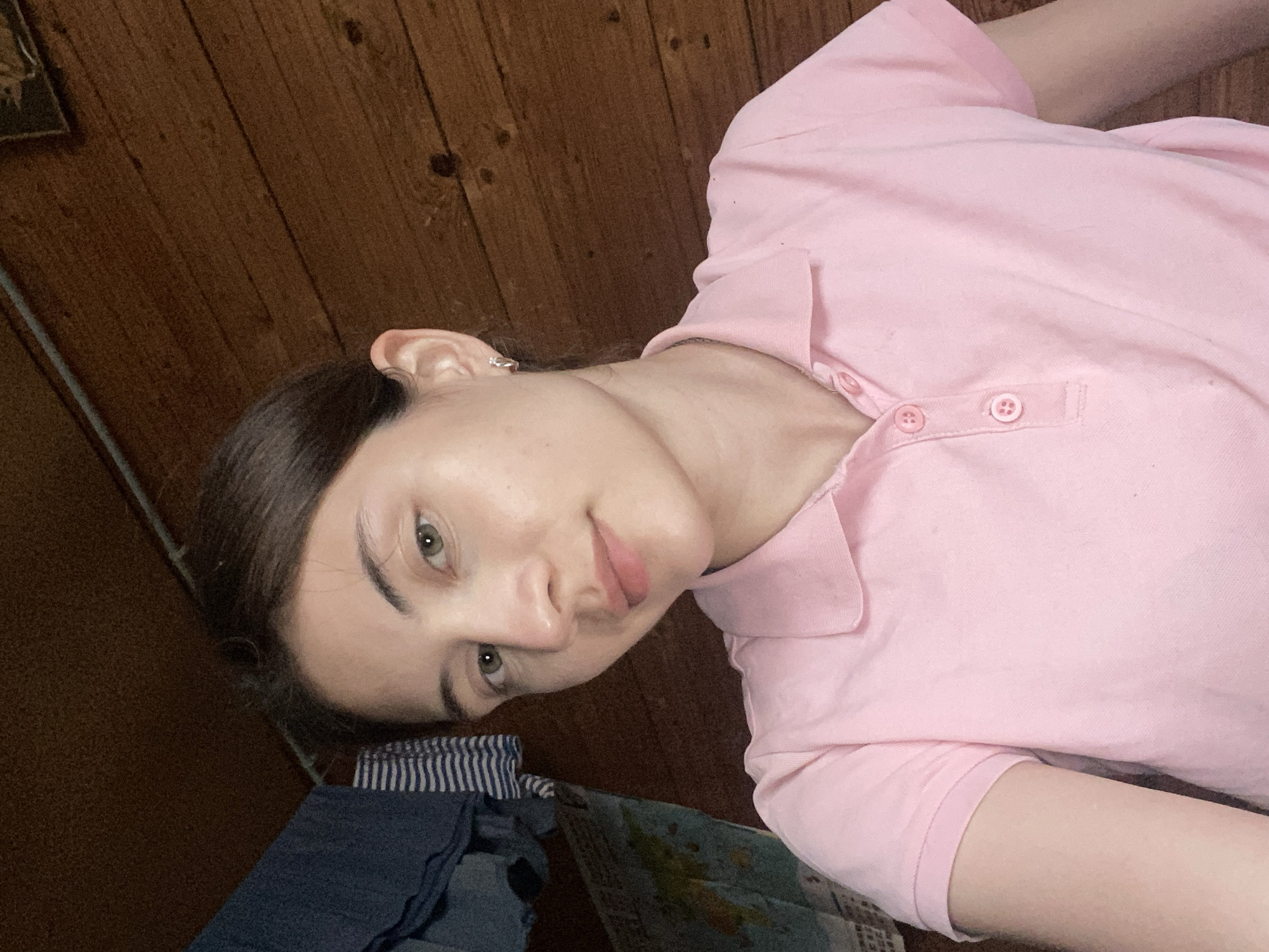 A young woman with light skin and dark hair pulled back is photographed from the chest up. She is wearing a pink polo shirt with two buttons at the collar. Her eyes are a light green, and she has a subtle smile. Her left ear is visible, adorned with a small silver earring. The background consists of a wooden wall and a section of what appears to be laundry hanging on a line to the left of the frame. A portion of a map is also visible on the left side. The lighting suggests an indoor environment. The image's metadata does not provide any information about its specific location, including Votkinsk, Russia.