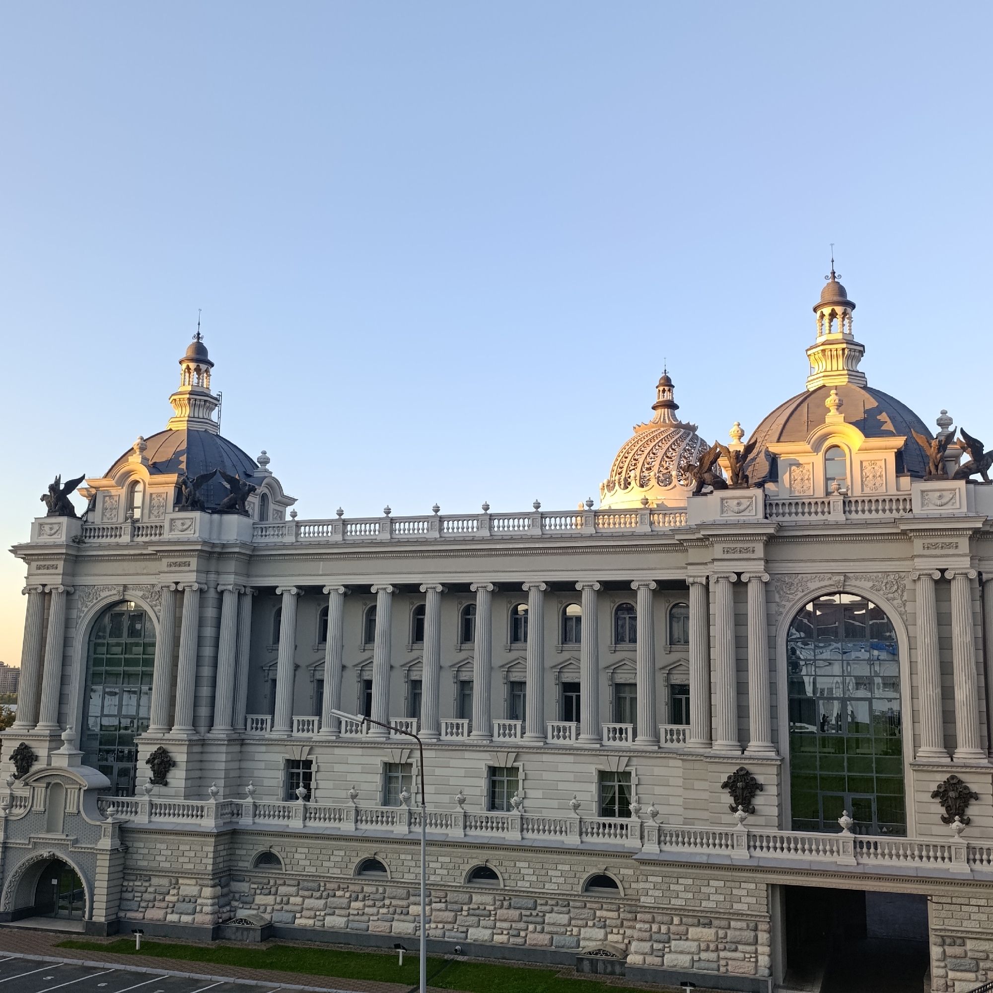 The image captures the Agricultural Palace in Kazan, Russia, a grand building with neoclassical architecture. The structure is characterized by its ornate facade, featuring numerous columns, intricate carvings, and two prominent domes. The building is illuminated by the soft glow of what appears to be a sunset or sunrise, casting warm light on the golden accents of the domes and the upper portions of the building. The sky is a clear, pale blue, indicating fair weather. In the foreground, a streetlamp stands prominently, and a section of a parking lot is visible, with faint white lines marking parking spaces. The building's lower levels are clad in stone, with arched entrances and windows. Several decorative sculptures of winged creatures adorn the upper sections of the building, above the colonnade. The overall scene depicts an architectural landmark under a serene sky.