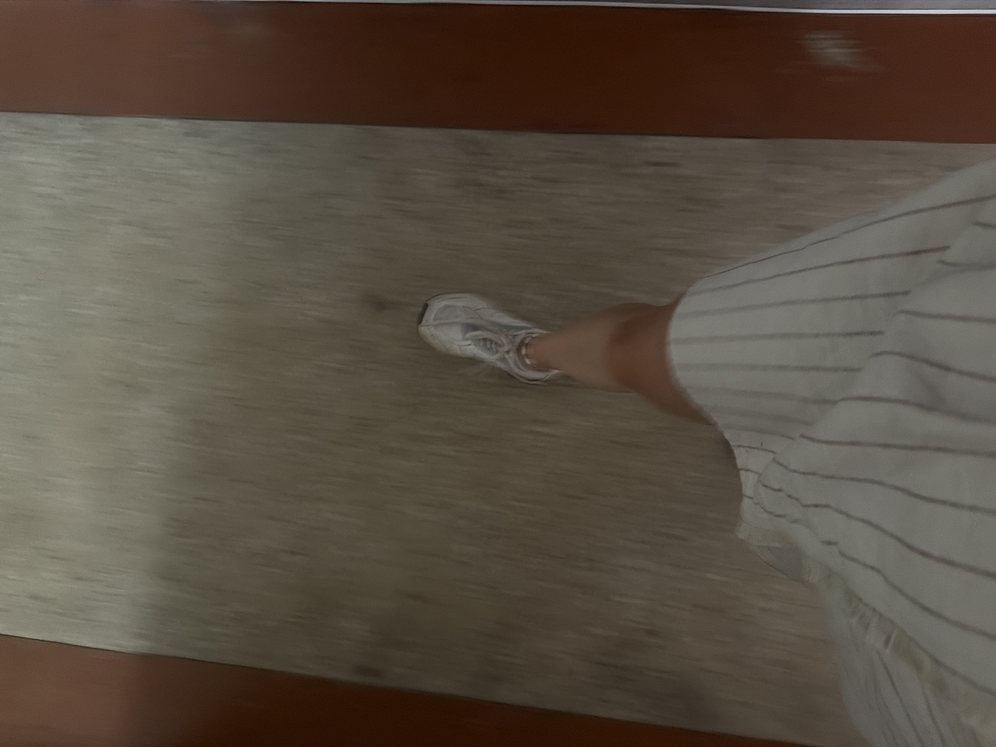 The image shows a person's legs and torso, with a white and grey striped dress and white sneakers, walking on a textured, light-colored floor. The floor appears to be an indoor walkway, possibly a corridor or hallway, with the sides of the walkway lined with reddish-brown panels. The motion blur suggests the person is moving. There is no visible indication of the location being in Chișinău, Moldova.